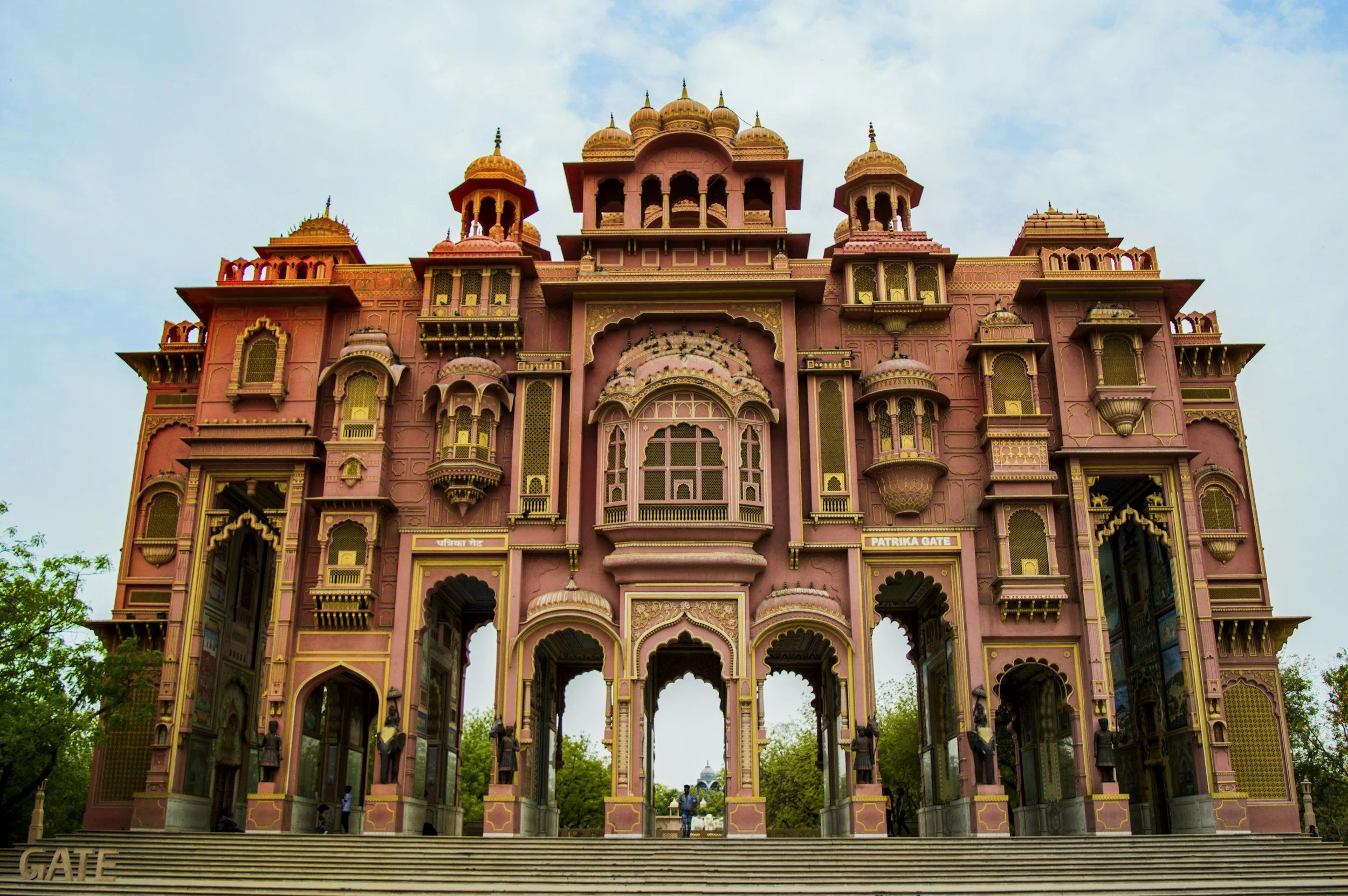 Front view of an ornate pink architectural gateway with arches, domes, and intricate gold decorations, labeled 'Patrika Gate', with a staircase leading up to it and trees surrounding.