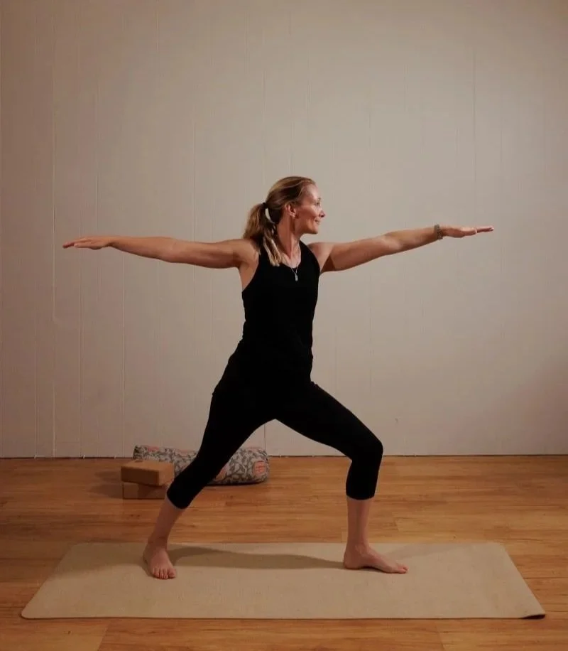 A woman practicing yoga in a warrior pose on a beige mat in a room with wooden flooring and a plain wall.