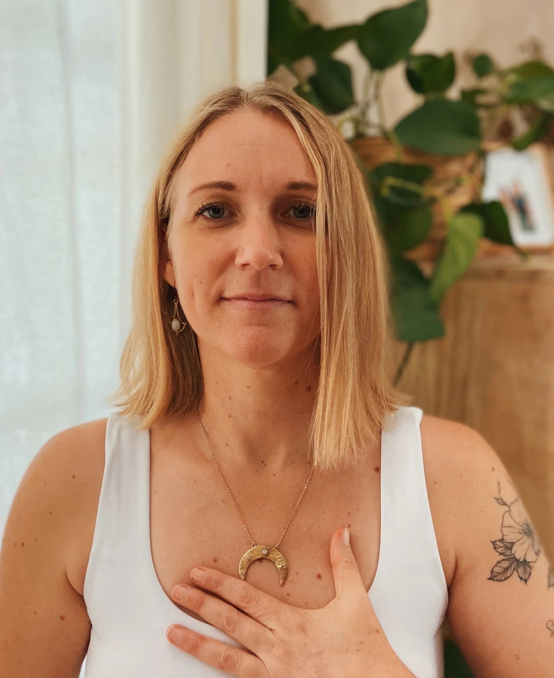 Lady with blonde hair with hand to her chest wearing a white vest