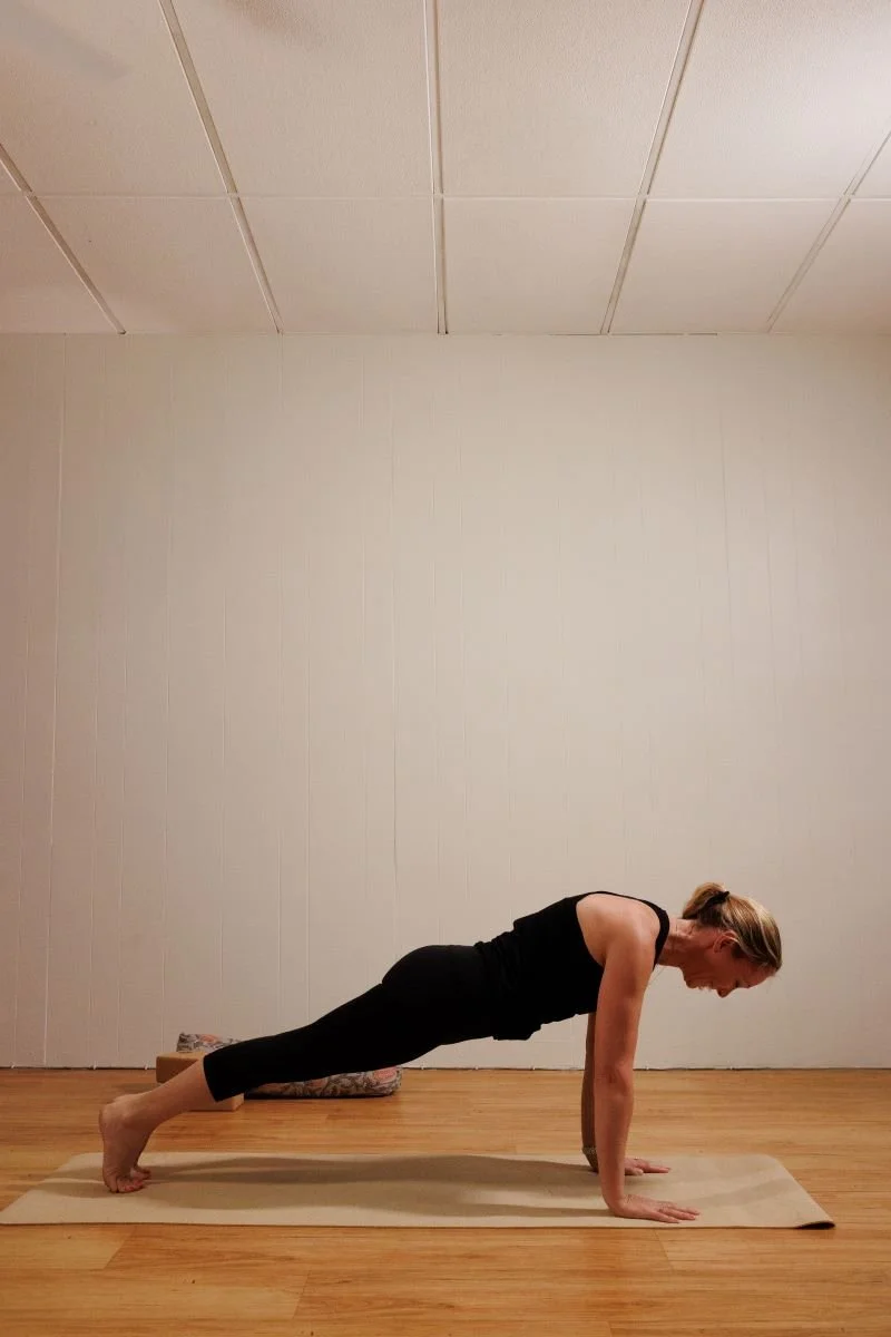 A woman in black workout clothes practicing yoga in a plank pose on a beige mat in a room with wooden floors and plain white walls.