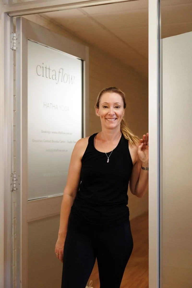A woman stands in a doorway, smiling, wearing a black sleeveless top and black pants, with a background that includes a sign for CittaFlow Hatha Yoga.