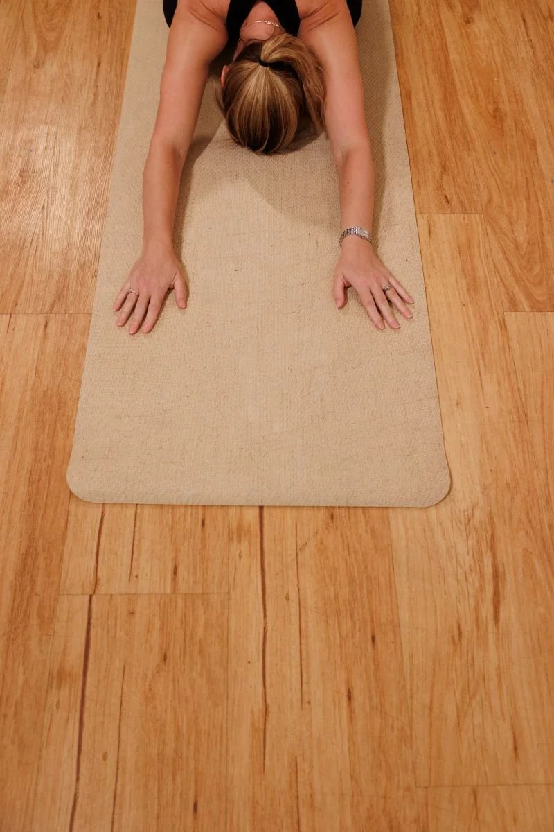 A woman practicing yoga or stretching on a beige mat on a hardwood floor, seen from above with arms extended forward.