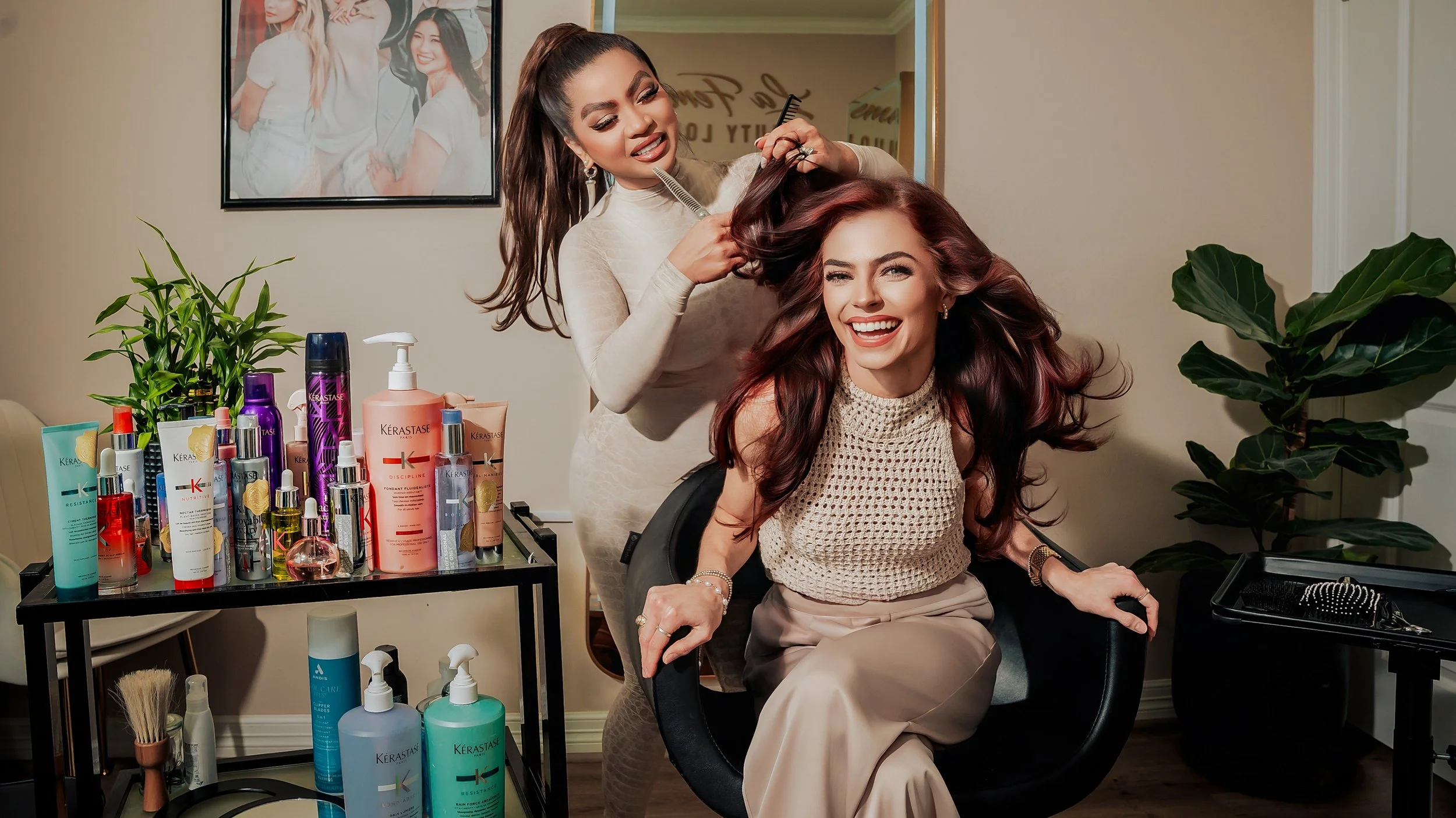 A woman sitting in a salon chair getting her hair styled by a stylist. The stylist is holding a section of the woman's long, wavy, auburn hair. The woman is smiling broadly. On the table in front of them, there is an assortment of hair care products,
