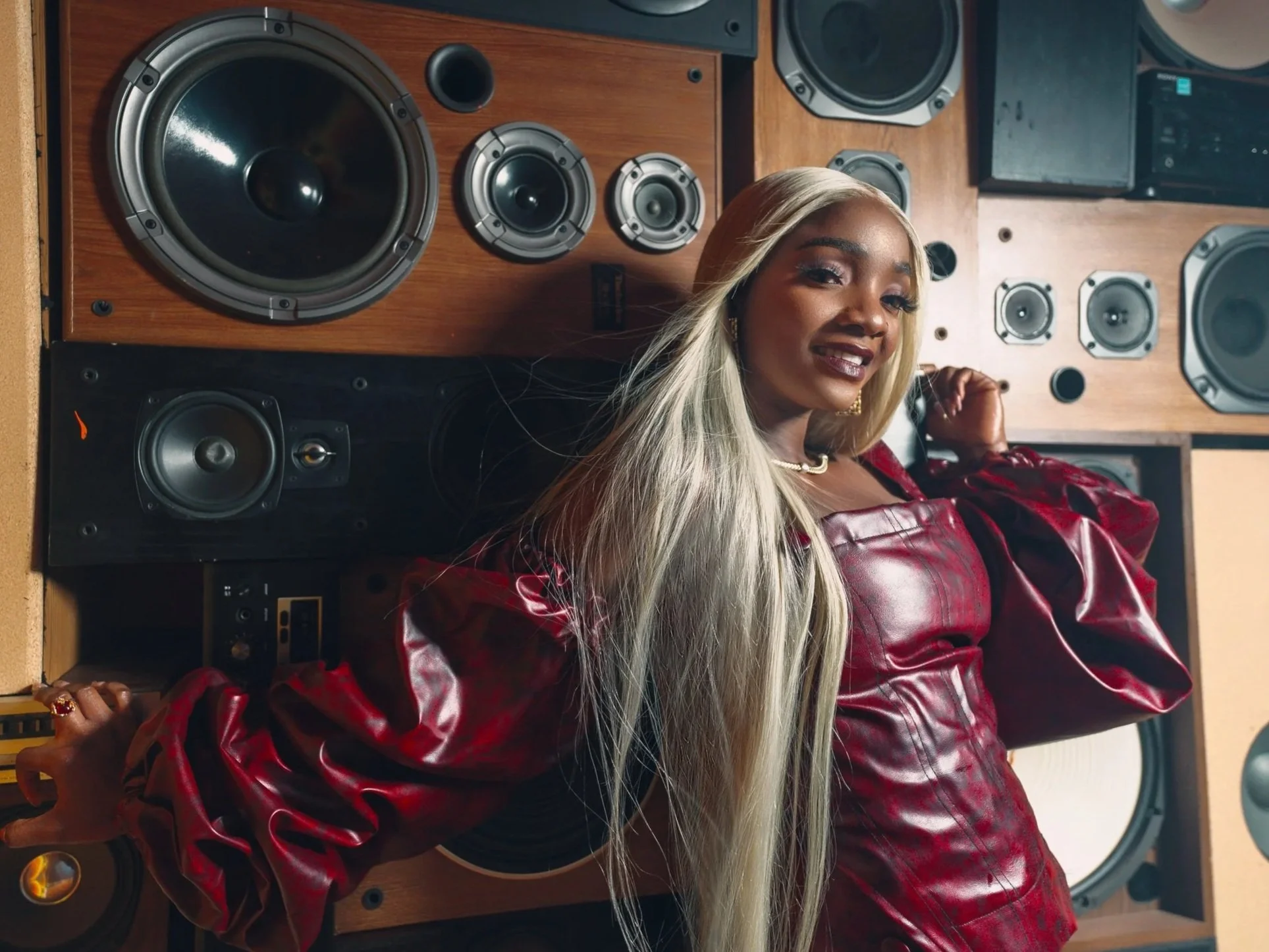 A woman with long blonde hair and a red leather jacket smiling and posing in front of large speakers in a music studio.
