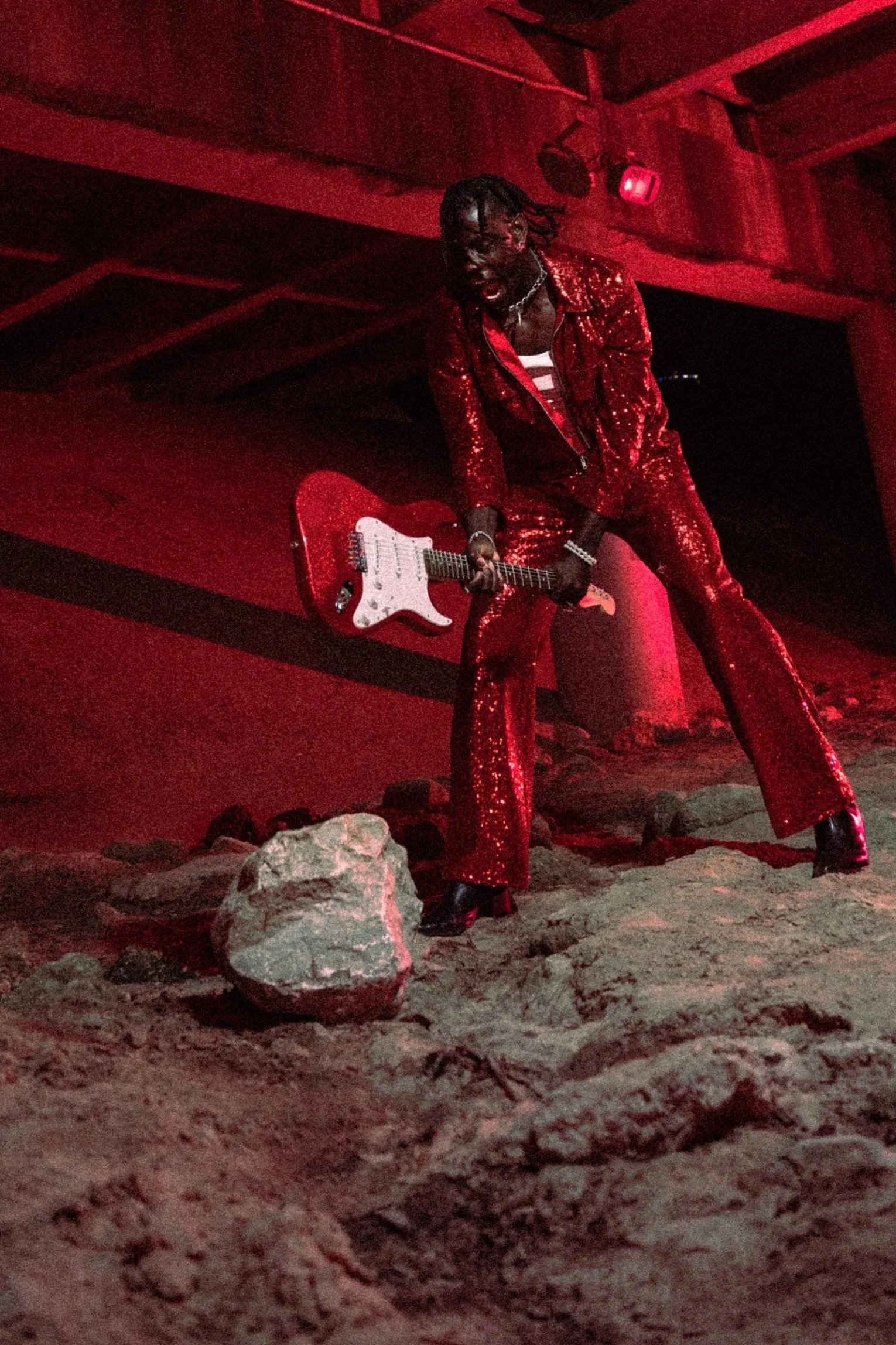 A person dressed in a shiny red suit holding a red and white electric guitar, standing on a rocky surface under a concrete structure, illuminated by red lighting.