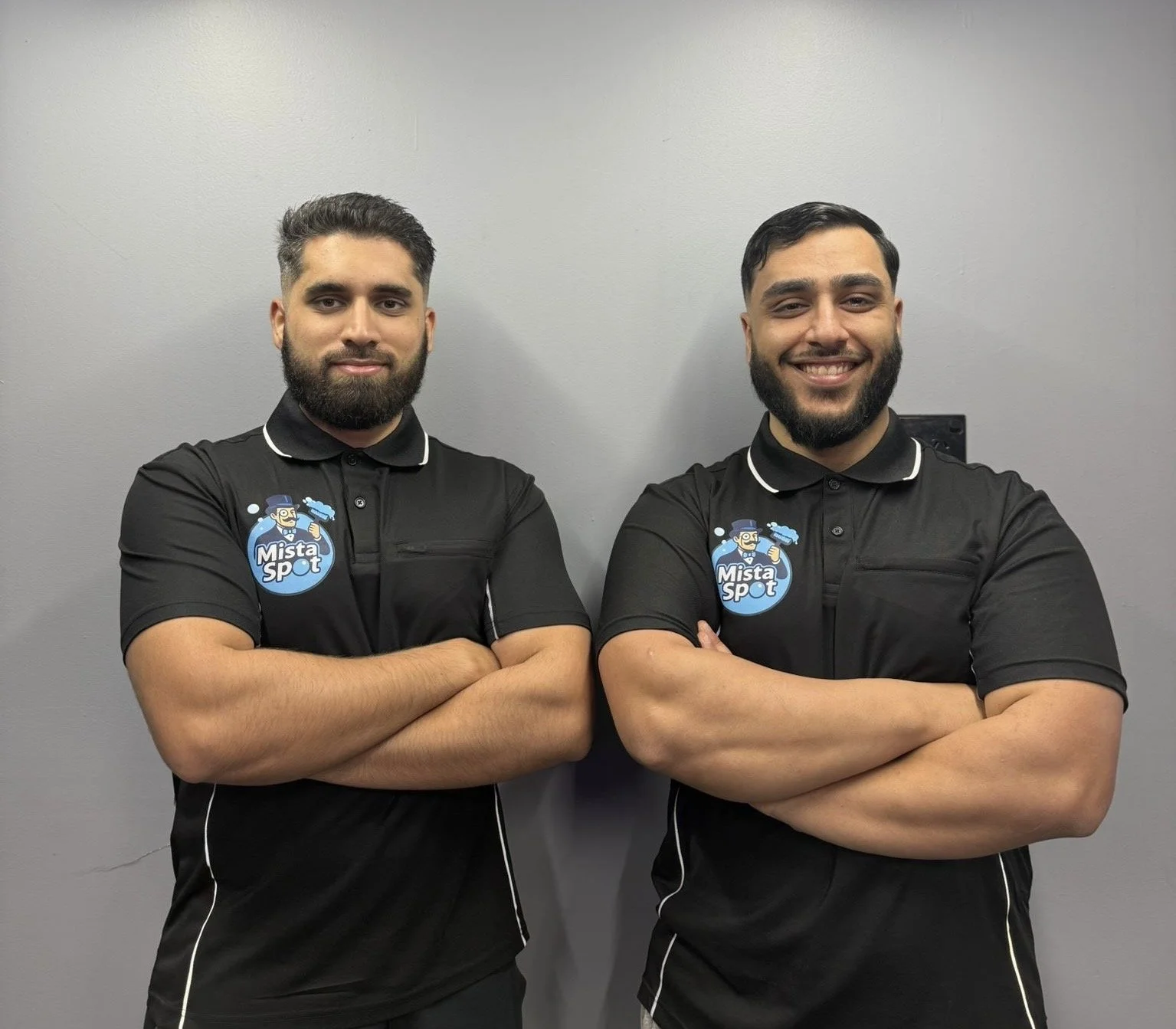 Two men with beards and dark hair wearing matching black polo shirts with 'Mista Spot' logo, standing with arms crossed against a gray wall.