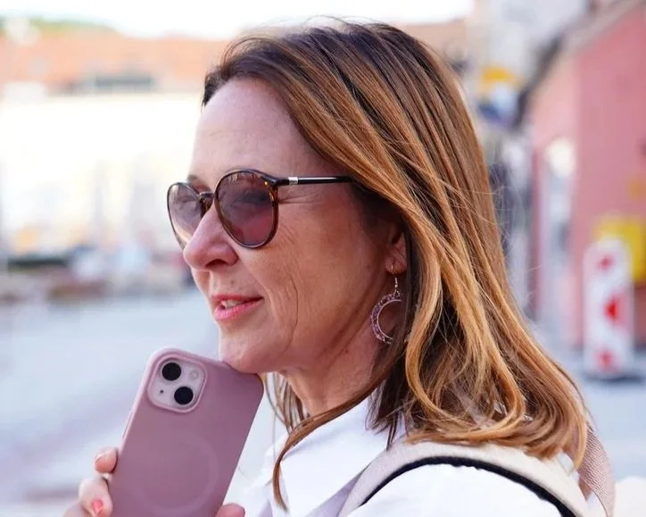 A woman with shoulder-length reddish-brown hair, sunglasses, and earrings, holds a smartphone near her face while walking outdoors in a city.