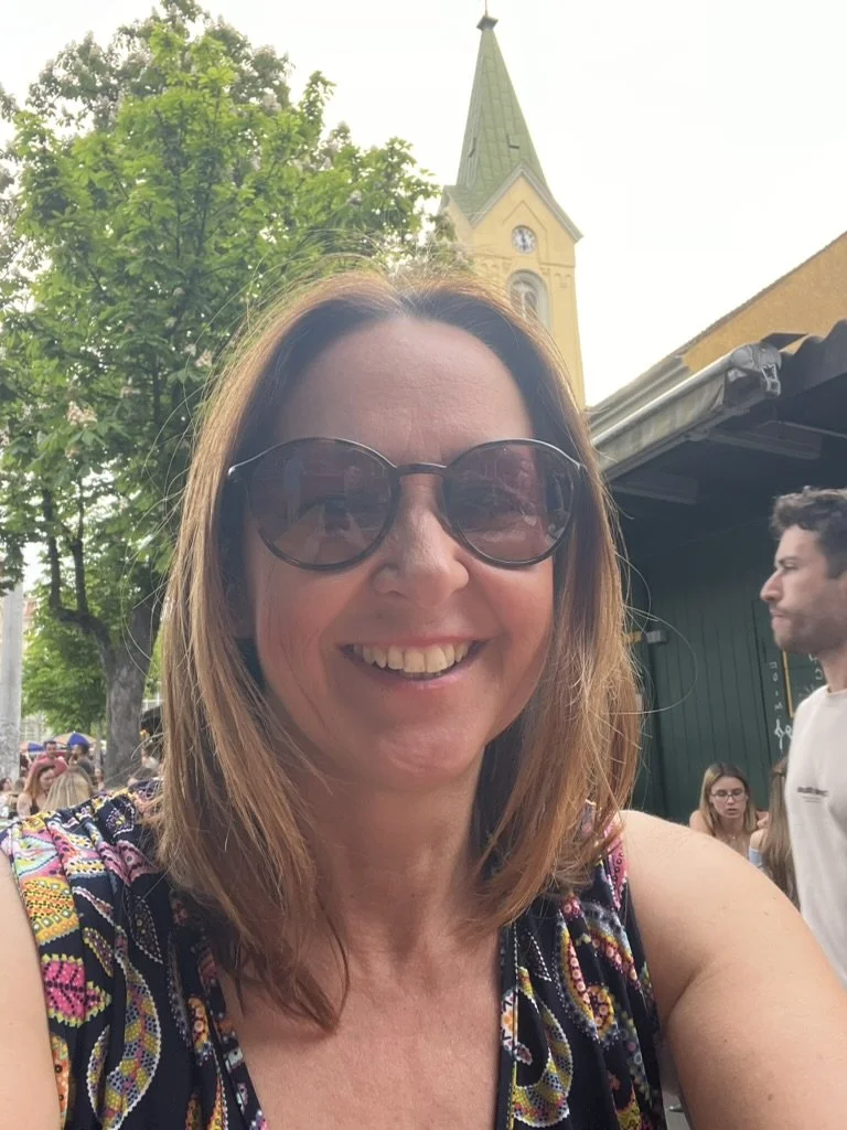 A woman with shoulder-length brown hair wearing large sunglasses and a colorful patterned top, smiling at the camera. In the background, there are trees, a church steeple, and other people at what appears to be an outdoor event.