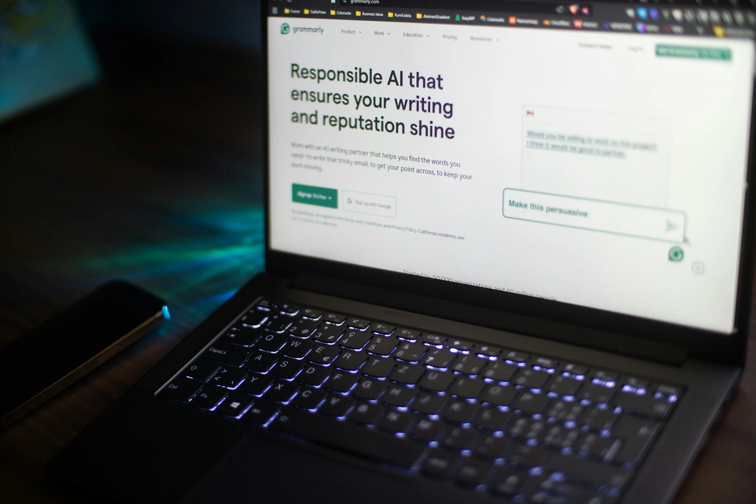 Laptop displaying Grammarly website with text promoting responsible AI for writing and reputation; keyboard illuminated, phone on the left, dark environment.