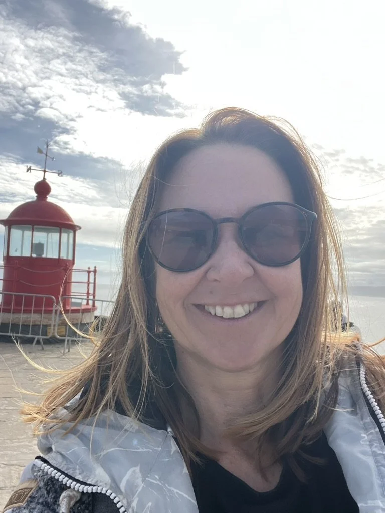 Smiling woman wearing sunglasses and a jacket standing on a beach near a red lighthouse with cloudy sky in the background.