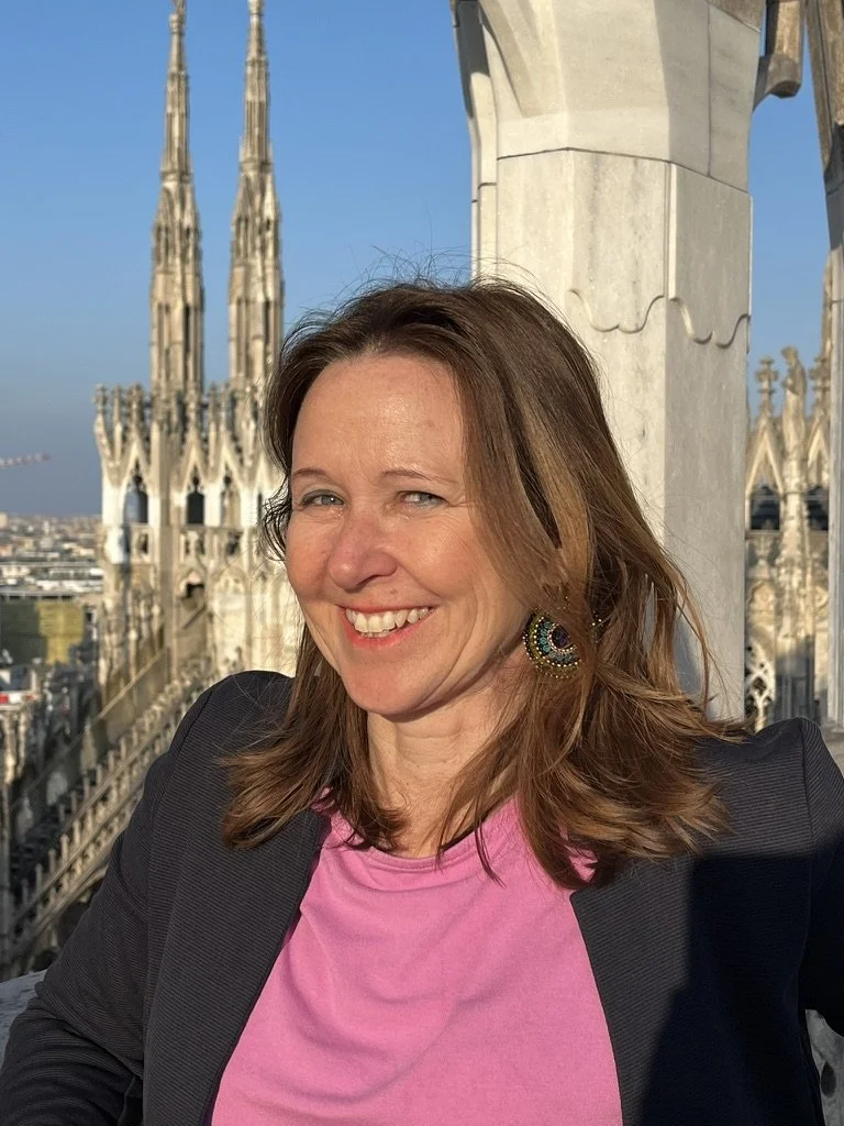 Woman with shoulder-length brown hair smiling outdoors on a rooftop with the city skyline and church spires in the background, wearing a pink top and dark blazer.