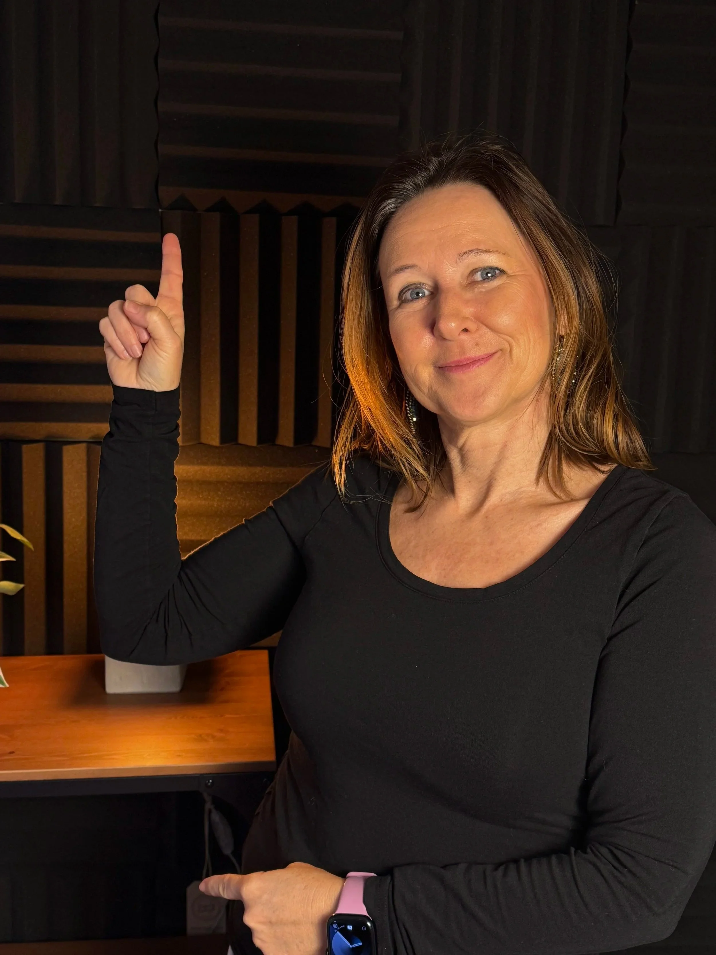 Woman in black shirt pointing upward with her index finger, sitting at a desk in a soundproof studio with foam panels.