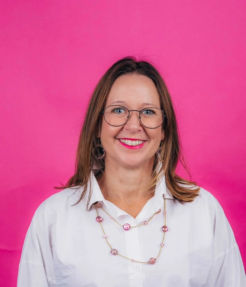 A woman with shoulder-length brown hair, wearing glasses, a white shirt, and a pink beaded necklace, smiling in front of a pink background.