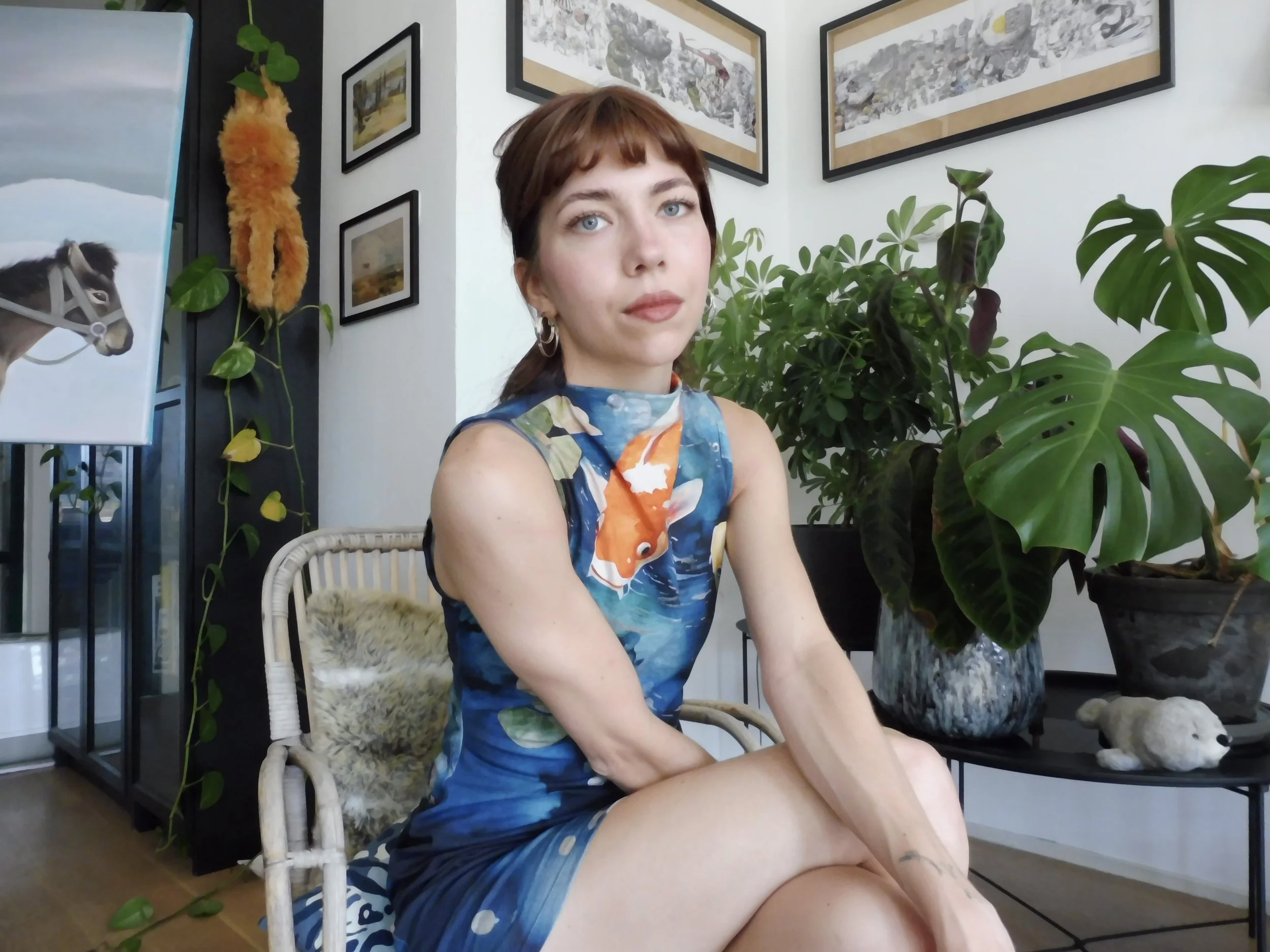 Een vrouw met kort bruin haar en blauwe ogen zit binnen, omringd door groene planten en kunstwerken aan de muur. Ze draagt een kleurrijk, visachtig patroon jurkje.