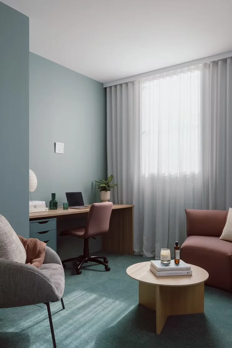 A cozy living room with a pastel green accent wall, sheer white curtains, a small wooden desk with a pink swivel chair, a gray armchair with a brown cushion, a pink sofa with a beige pillow, and a round coffee table with books, candles, and bottles.