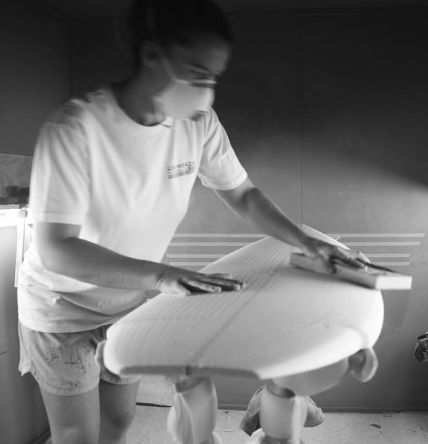 A woman in a t-shirt and shorts, wearing a face mask, is sanding or polishing a surfboard. Aitken Surfboards