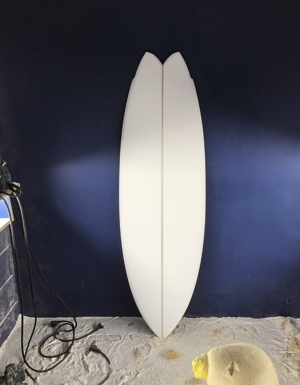 A white surfboard leaning against a dark blue wall in a sandy area. Aitken Surfboards