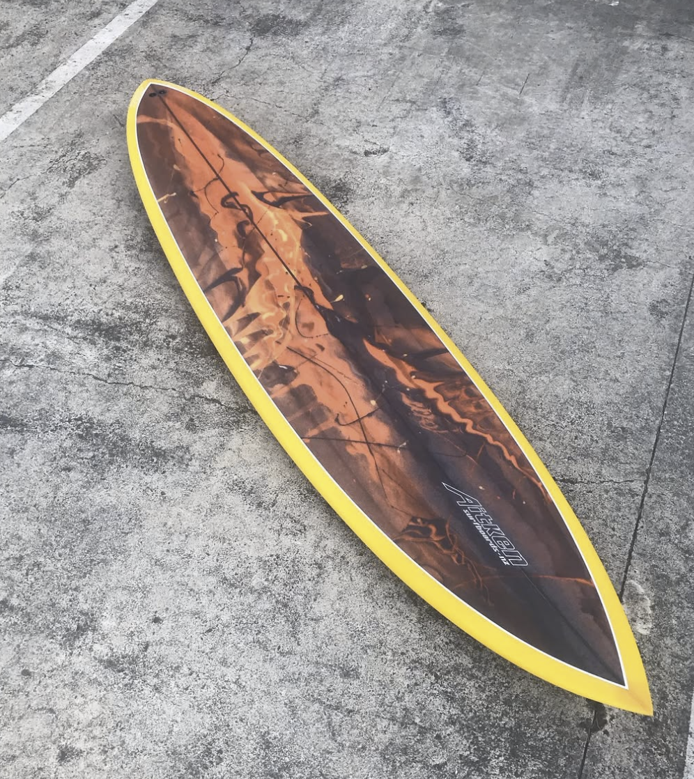 A yellow-edged surfboard with a fiery, flame-like design on top, lying on a concrete surface. Aitken Surfboards