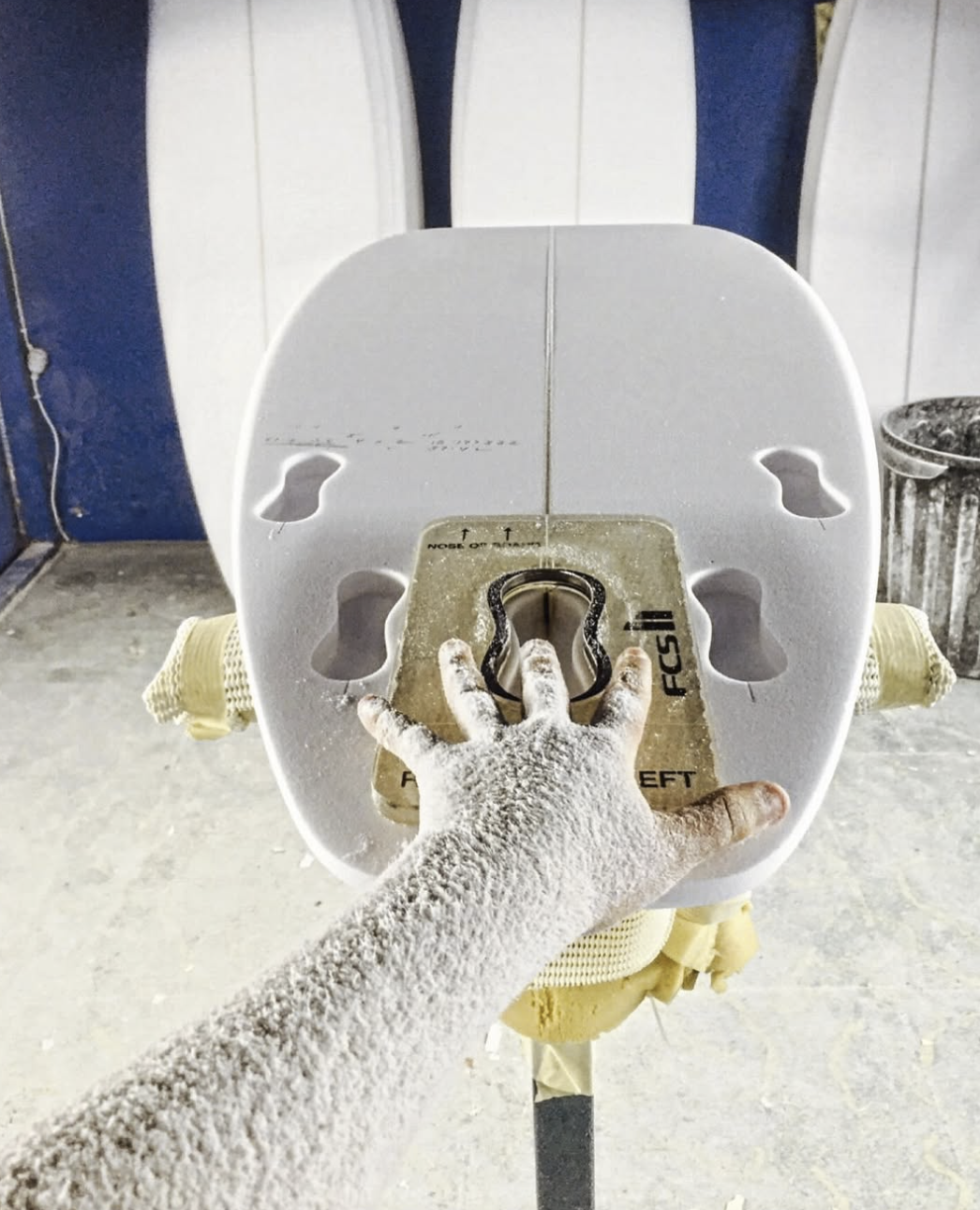 Close-up of a hand covered in white powder, adjusting a helmet on a yellow surfboard with foot holes in a workshop or storage area. Aitken Surfboards