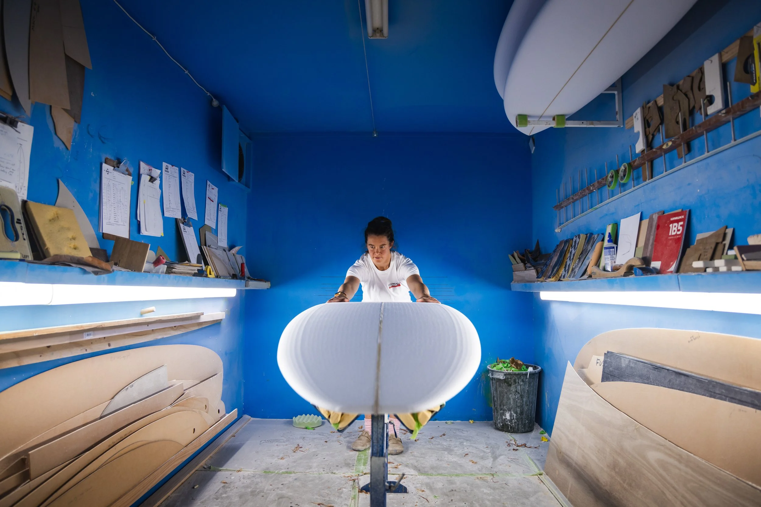 A person working on shaping a surfboard inside a blue workshop room, with surfboards, tools, and papers on shelves around. Aitken Surfboards