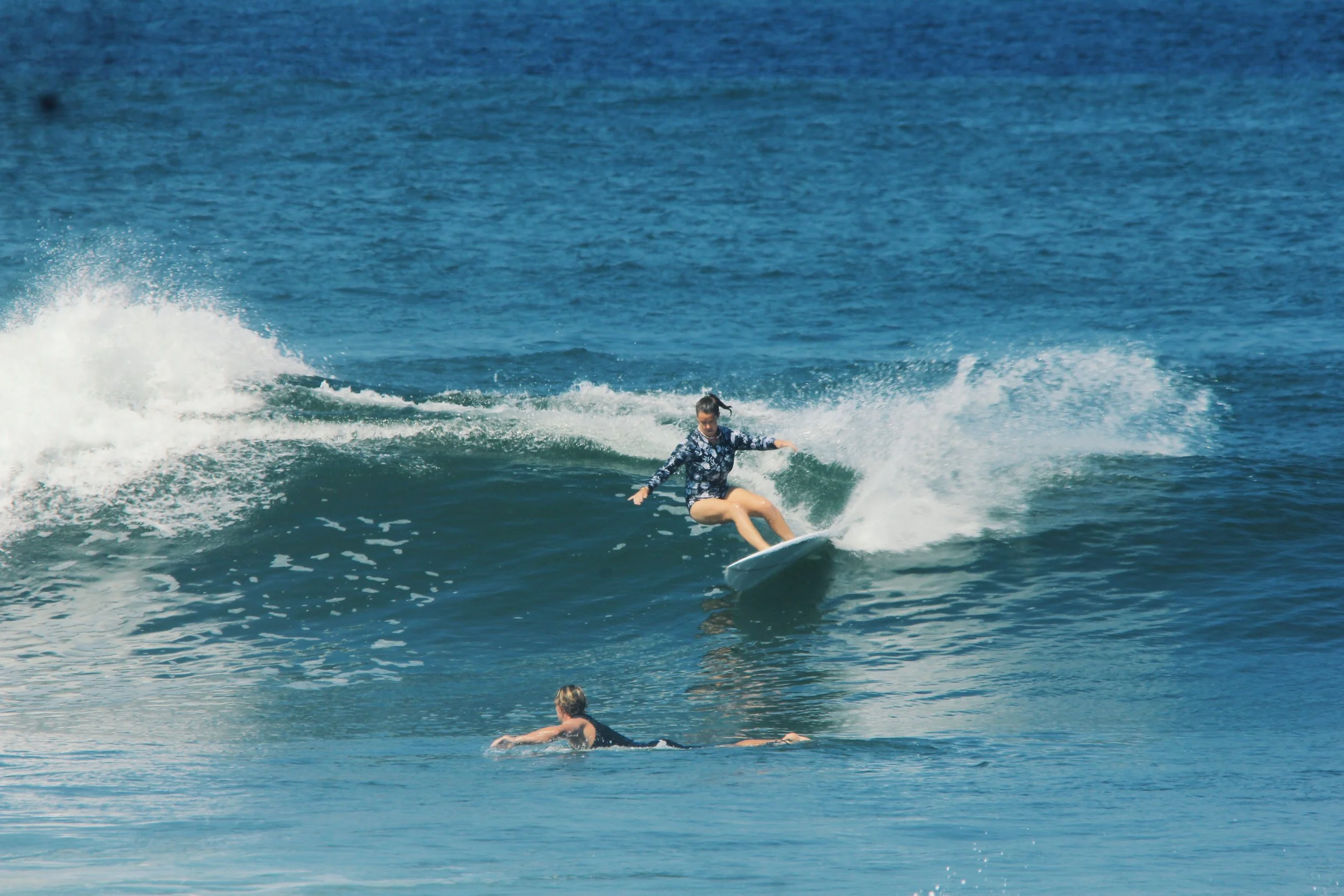 A person surfing on a wave in the ocean while another individual floats nearby. Aitken Surfboards