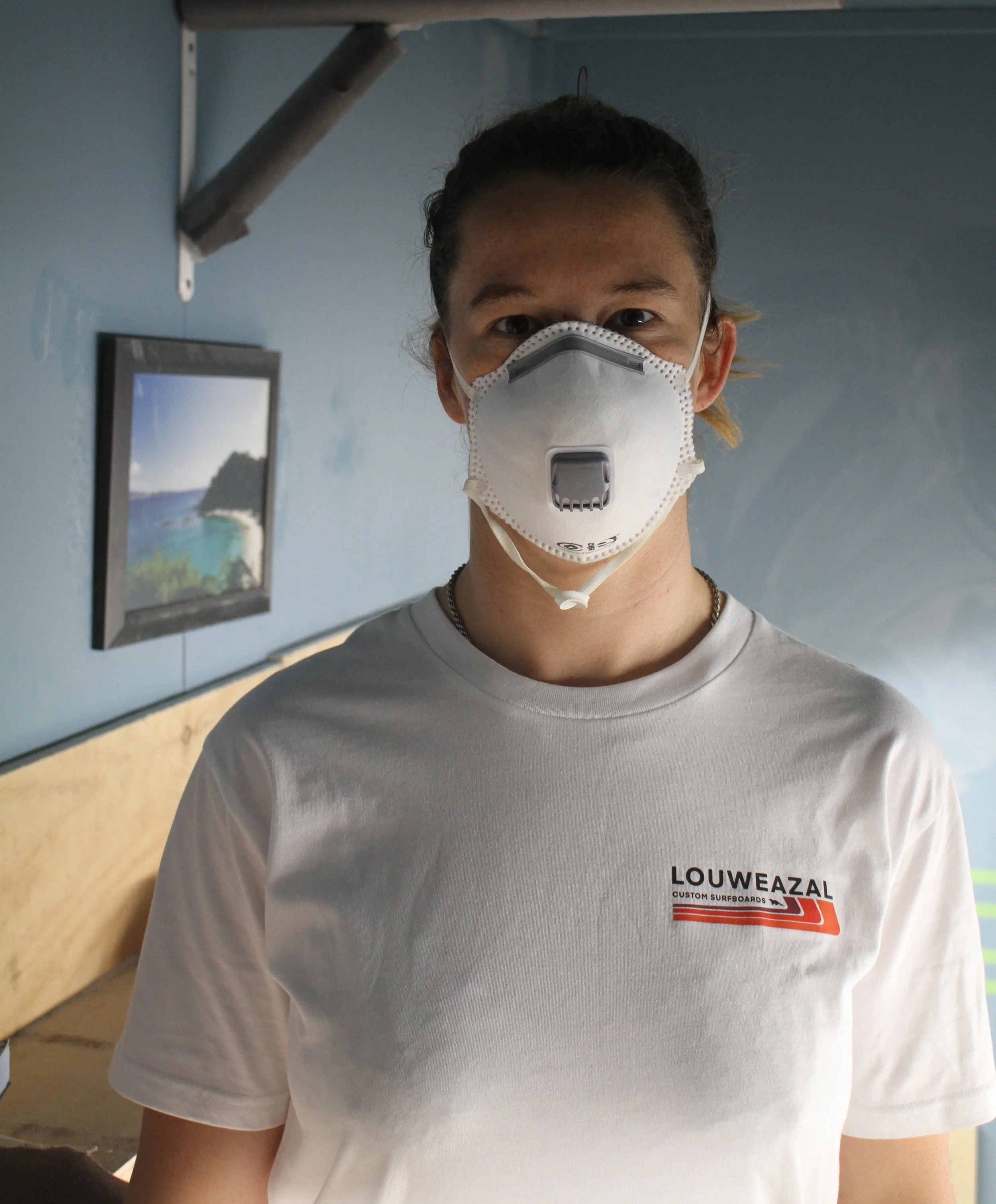Person wearing a white T-shirt with 'LOUWEAZAL' logo and a protective face mask, standing indoors with a blue wall and a framed picture in the background. Aitken Surfboards
