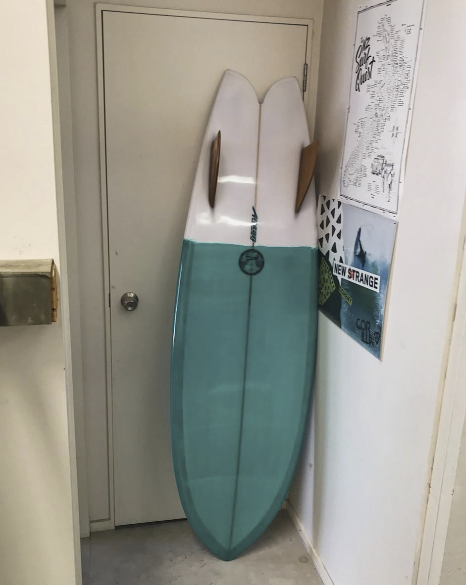 A surfboard with a half white, half teal design leaning against a door, with wooden fins attached to the top side of the surfboard. Aitken Surfboards