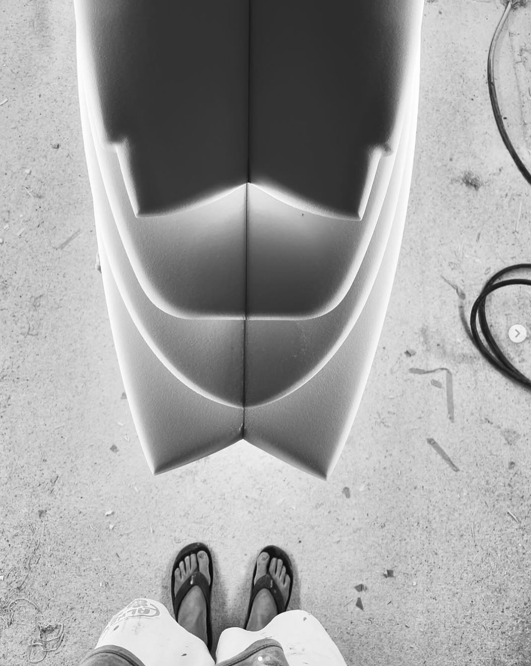 From a top-down perspective, a person is wearing flip-flops and shorts, standing on a textured concrete surface. In front of them is a surfboard with fin slots visible at the top of the image. Aitken Surfboards