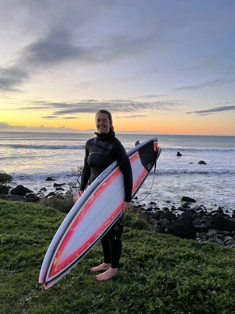 Person in wetsuit holding surfboard on grassy area near rocky beach at sunset. Aitken Surfboards