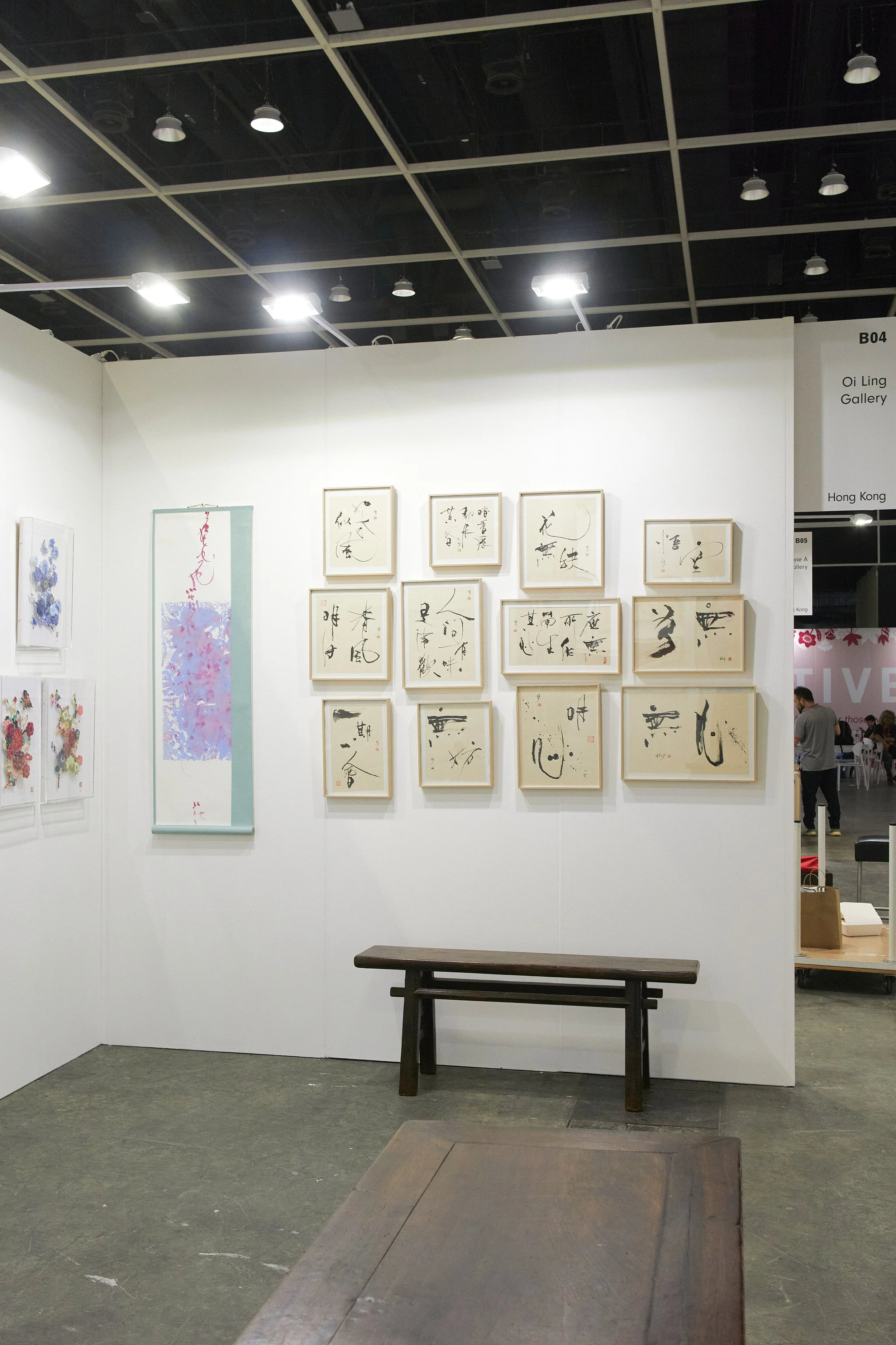 Affordable Art Fair 2025: Art gallery display featuring framed calligraphy and floral paintings on white walls, with a small wooden bench below.