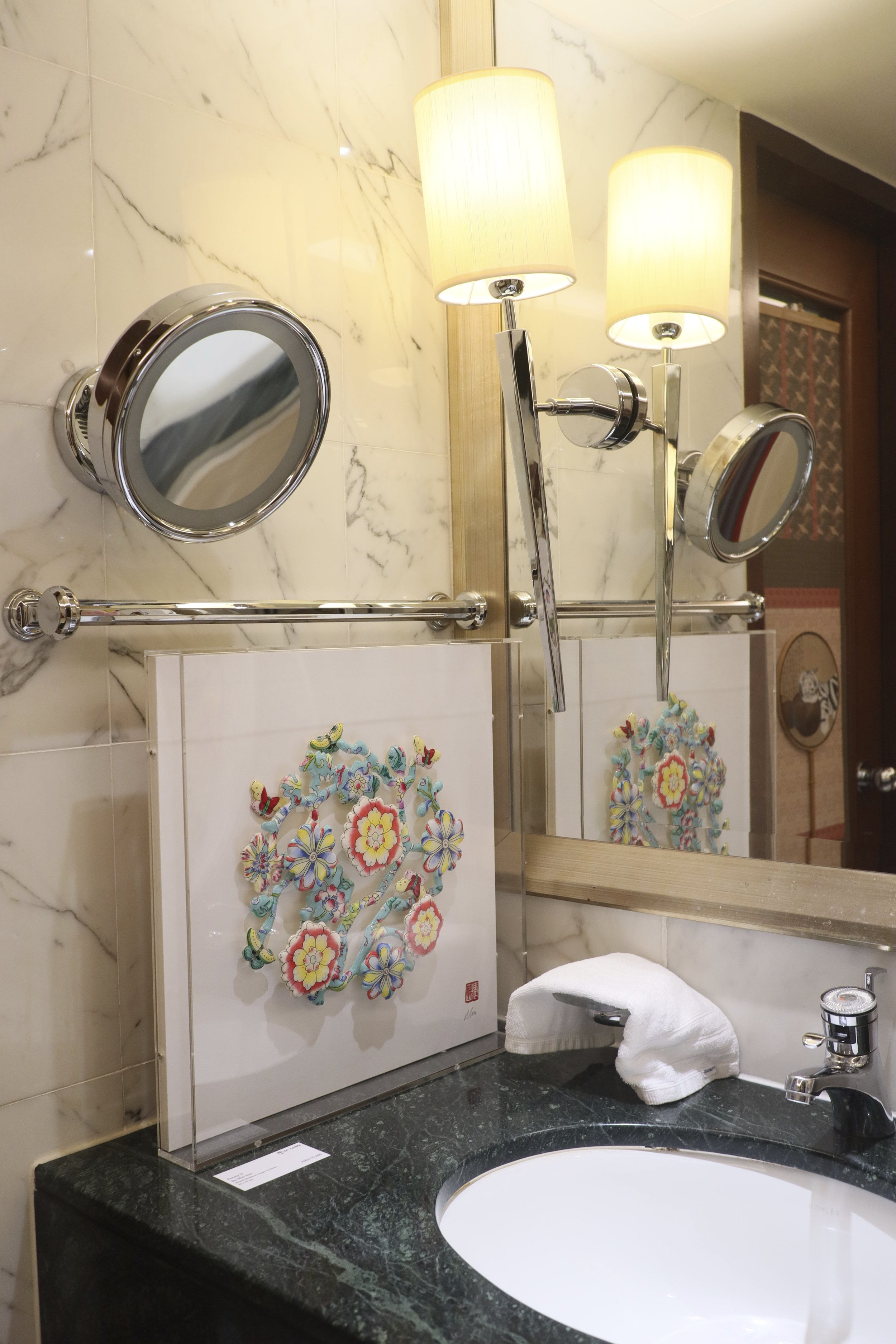 Whatz Art Fair 2025: A bathroom counter with a white sink, a towel, and artwork featuring colorful floral and butterfly designs, with a mirror and wall-mounted lights reflected in the mirror.