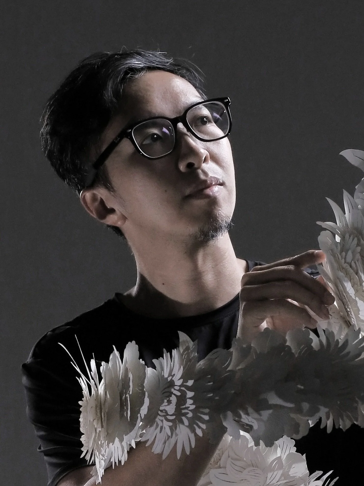 Joe Wong 黃文翰：A man with glasses carefully arranges a long, flowing chain of white artificial flowers against a dark background.