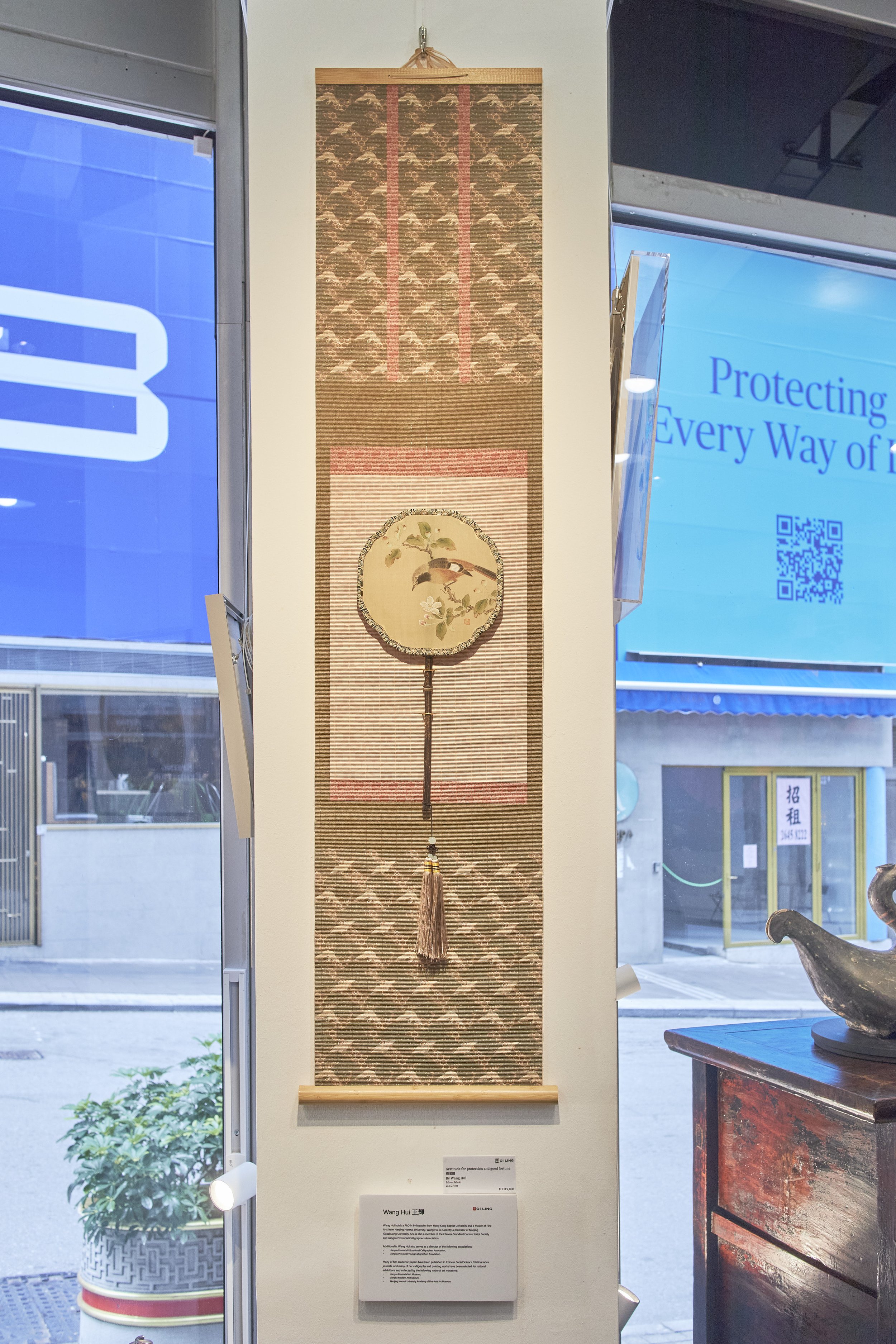 A vertical Asian hanging scroll with a painting of a small bird on a branch with blossoms, featuring a woven round fan with a bird and flowers motif, hanging from a beige and pink background.
