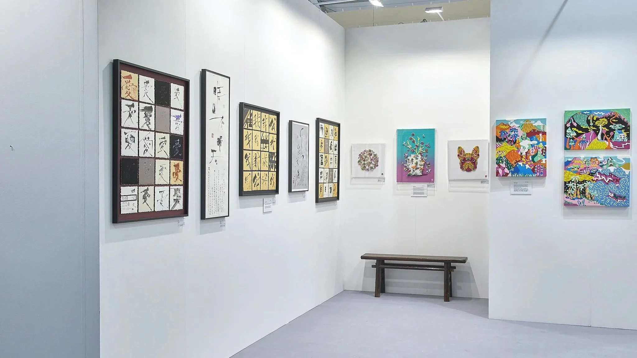Art Central 2025: Art gallery with various colorful artworks on white walls, including traditional calligraphy, intricate designs, and vibrant illustrations, with a wooden bench in the corner.