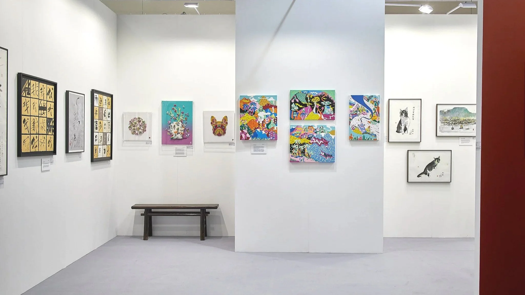 Art Central 2025: Art gallery with white walls displaying colorful paintings and illustrations, a wooden bench against the wall, and a red wall partially visible on the right.