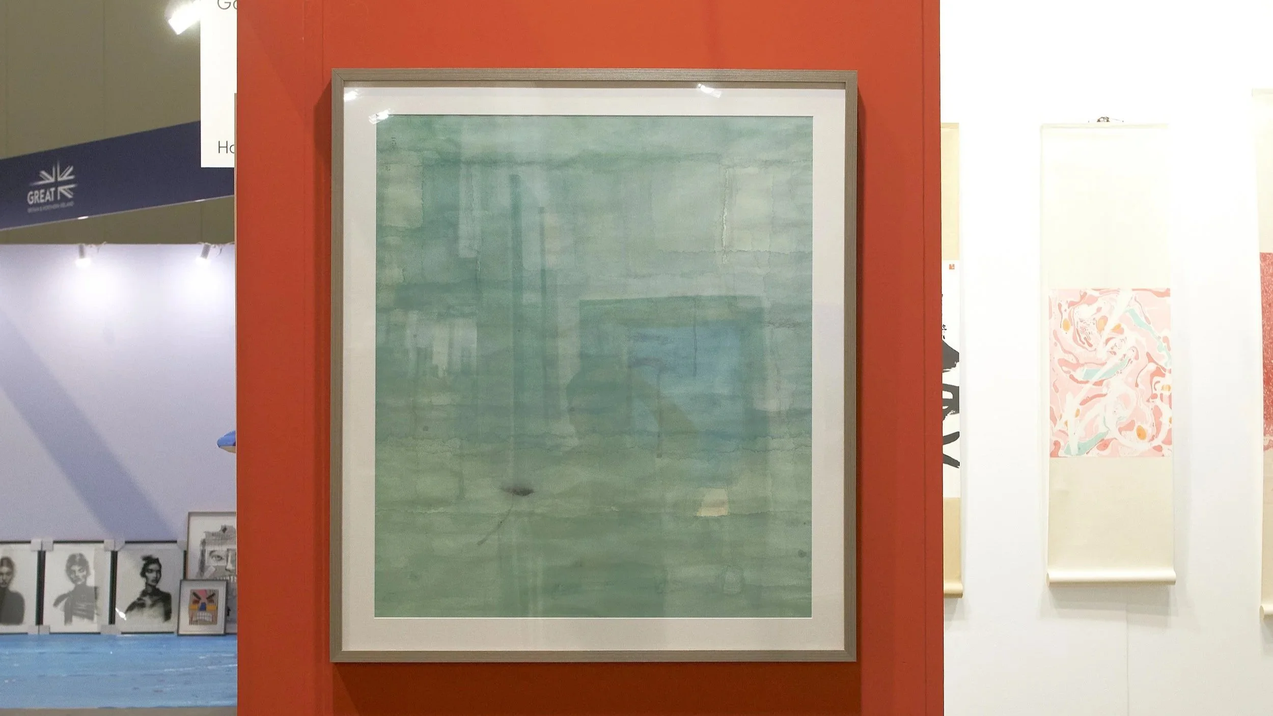 Affordable Art Fair 2025: A framed abstract painting with shades of green hangs on a red wall at an art gallery. In the background, there are white walls with other artworks, including a roll of patterned paper and photographs of people.
