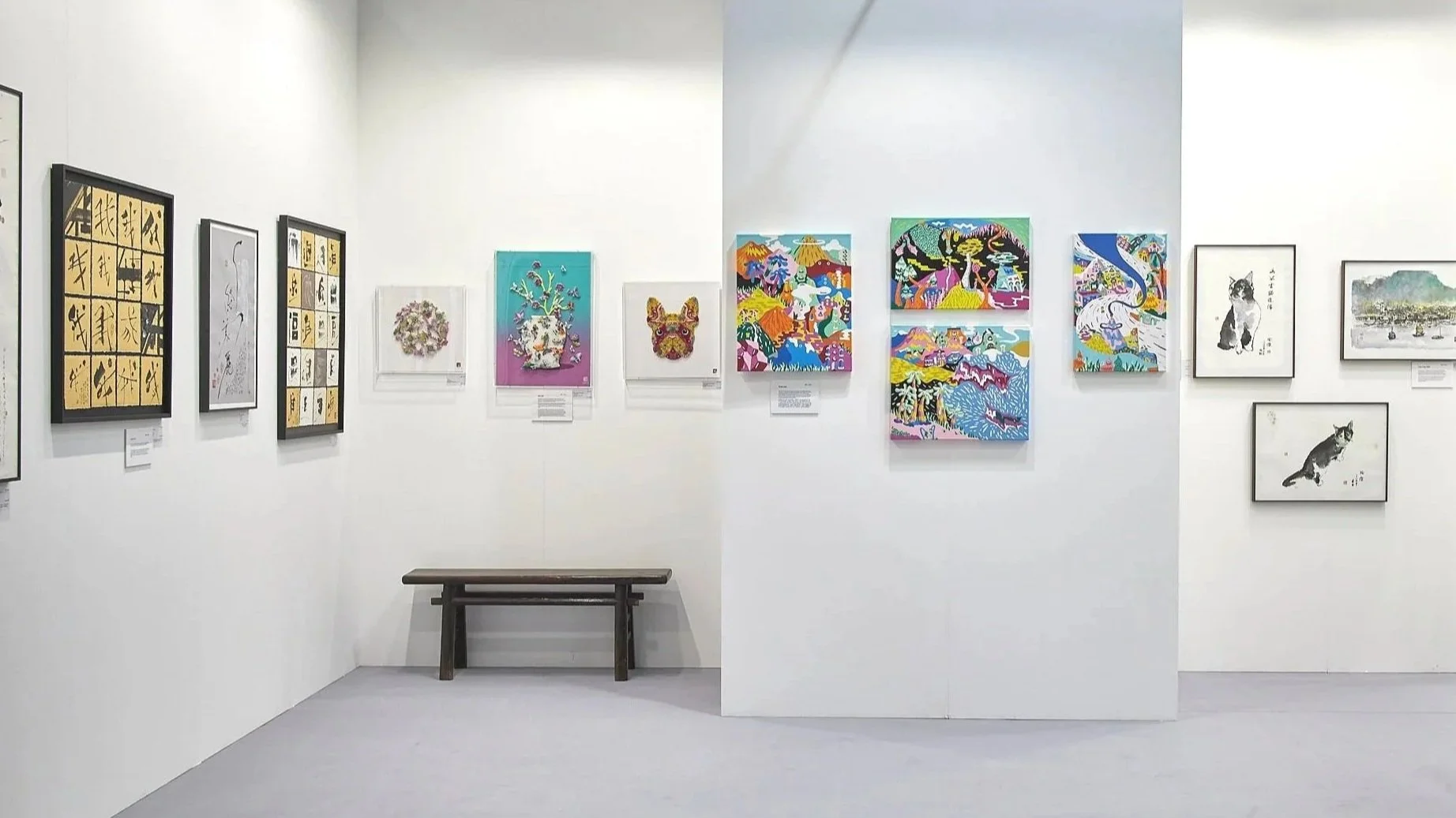 Art gallery with white walls displaying various colorful paintings and illustrations, with a wooden bench in the center.