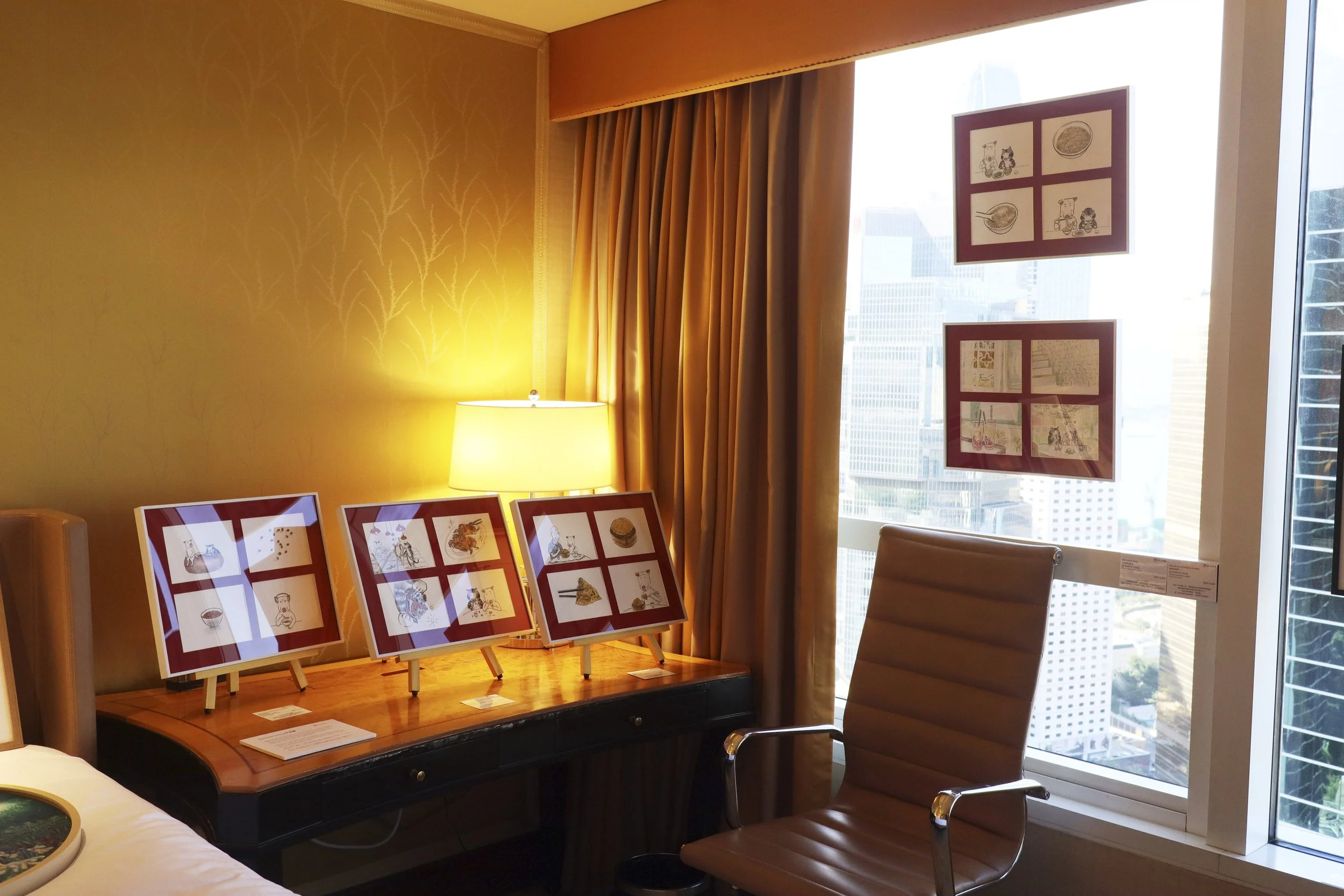 Whatz Art Fair 2025: Hotel room with a wooden desk displaying framed artwork, a beige leather chair, a bedside lamp, golden yellow curtains, and cityscape view through a large window.