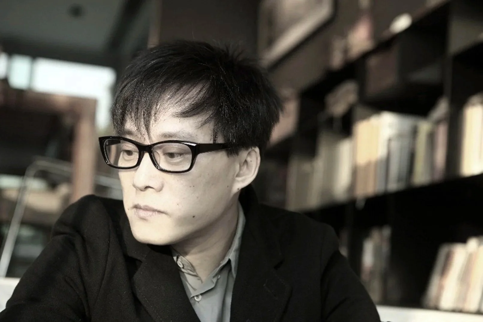 Lin Xi 林夕: A young man with black hair, black glasses, wearing a dark blazer and a light-colored shirt, sitting in a library or bookstore with shelves of books in the background.
