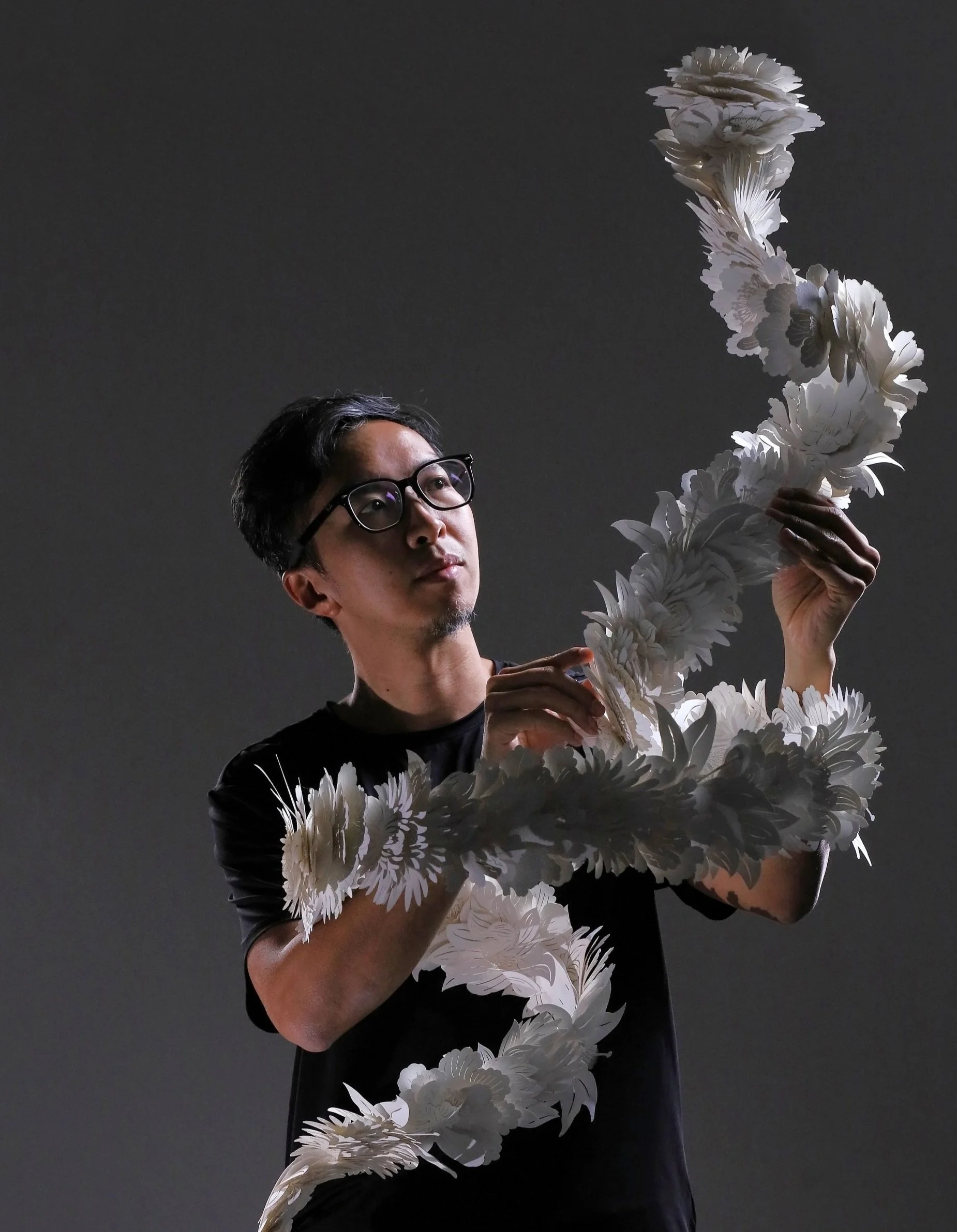Joe Wong 黃文翰：A man with black hair and glasses arranges a large swirl of white paper flowers against a dark background.
