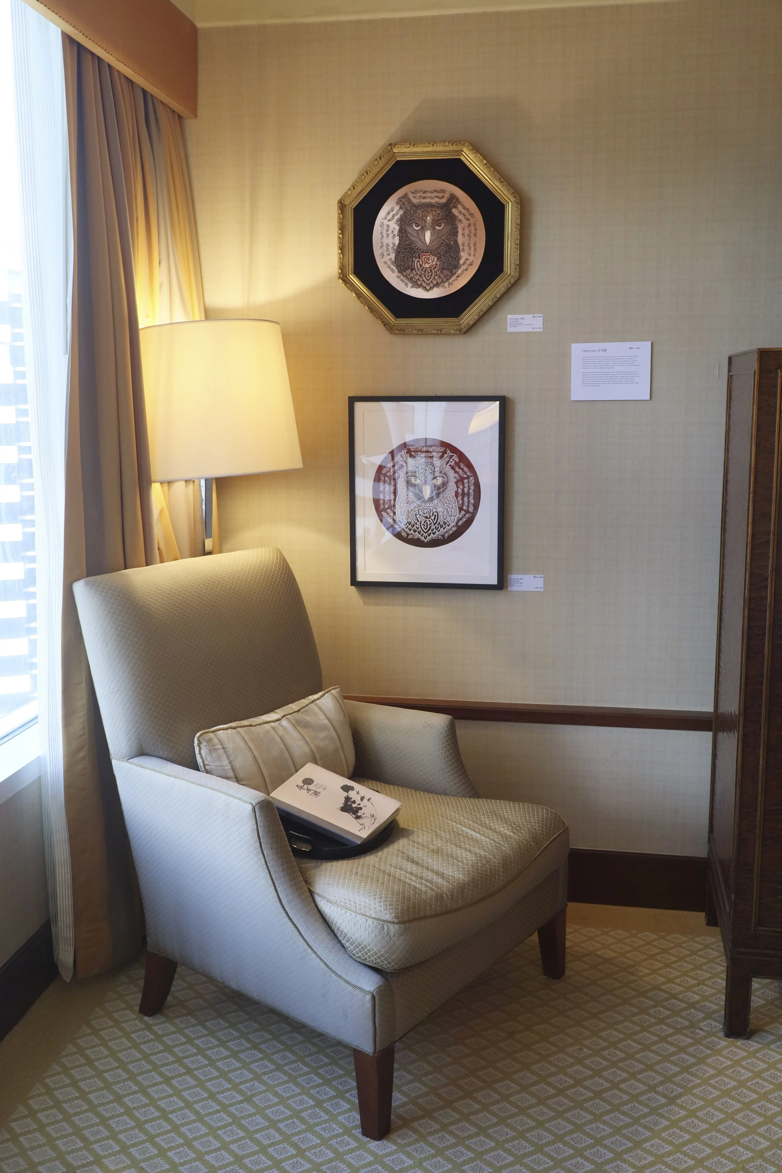 Whatz Art Fair 2025: A cozy corner in a hotel room featuring a beige armchair with a small pillow, a standing lamp, and framed artwork on the wall, with curtains and a window on the left side.