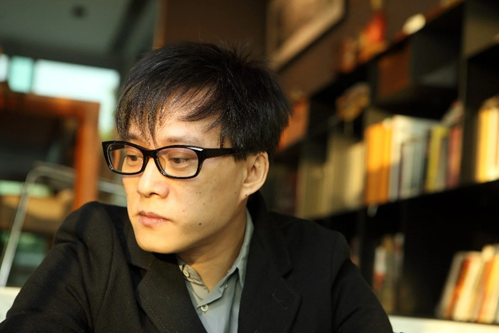 Lin Xi 林夕:  Young man with black hair and glasses sitting in a library, looking down, with bookshelves in the background.