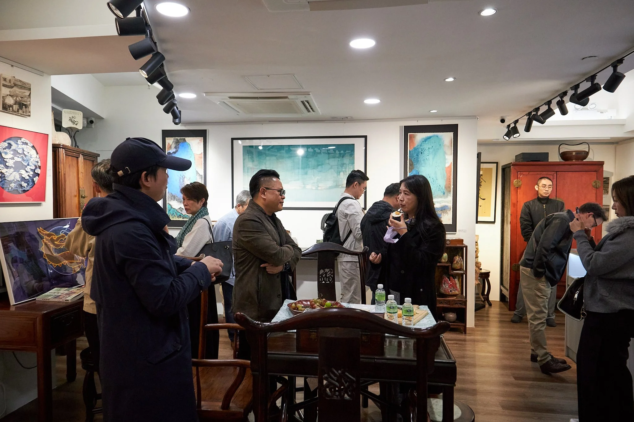 People attending an art gallery, viewing paintings and sculptures, some engaging in conversation, with artworks displayed on walls and furniture in the background.