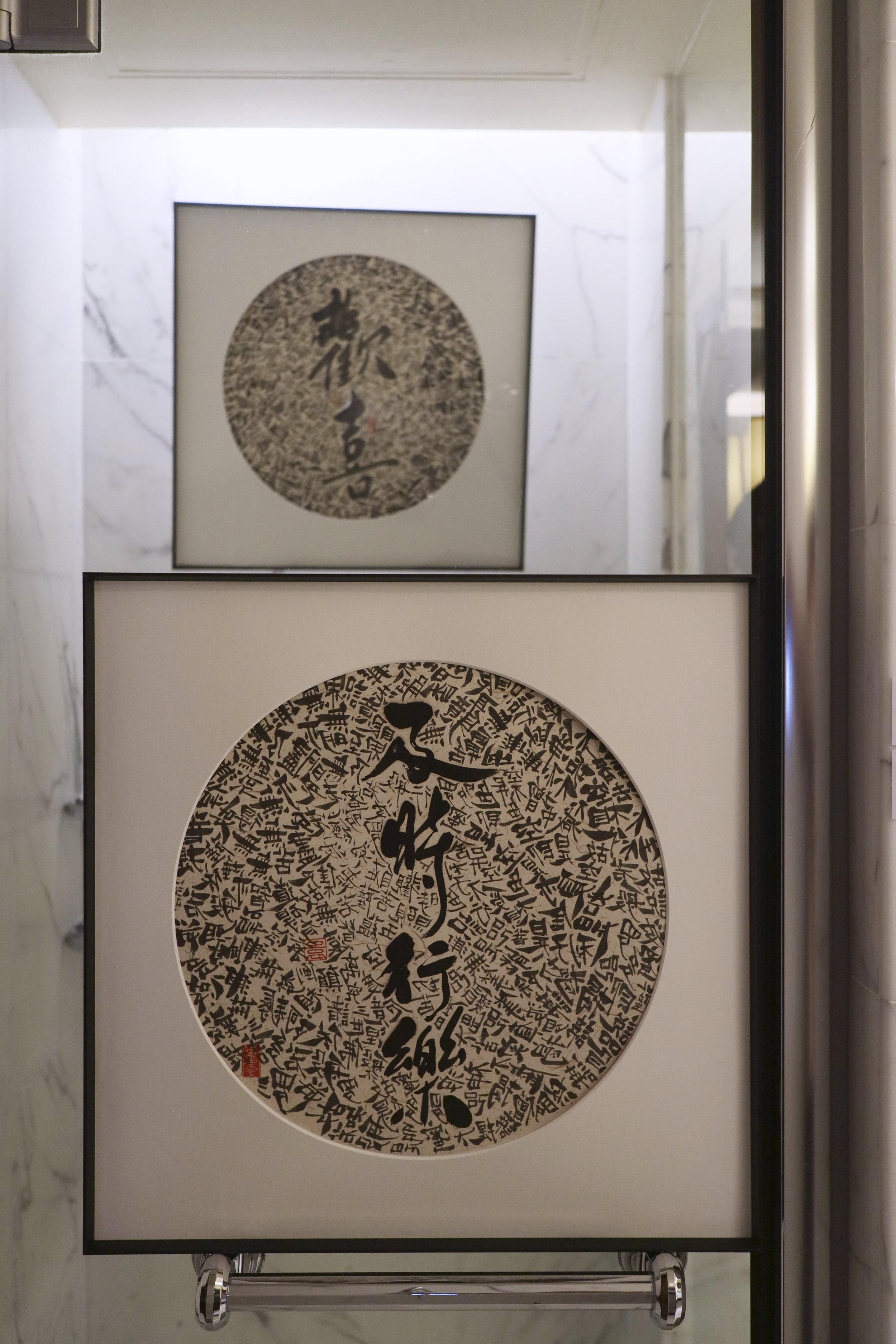 Two framed pieces of Asian calligraphy art, one on top of the other, hanging on a marble wall. The larger piece at the bottom has black calligraphy characters in a circular design with red stamps. The smaller piece at the top has a single calligraphy