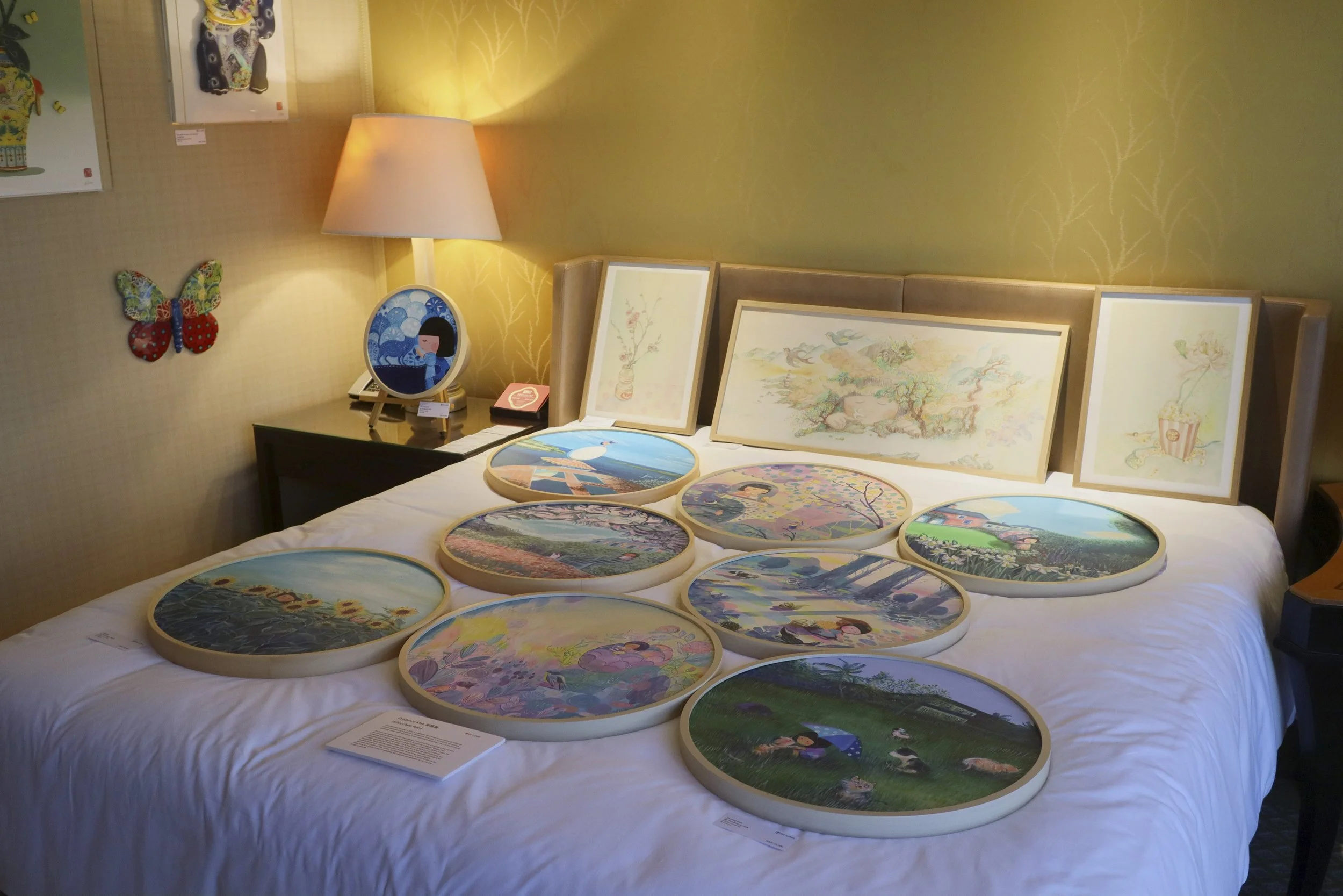 A bed with colorful embroidered embroidered artworks placed on it. The headboard has framed pastel illustrations. To the left, there's a bedside table with a lamp, a framed picture, and a small pink box. On the wall above the bedside table is a butte