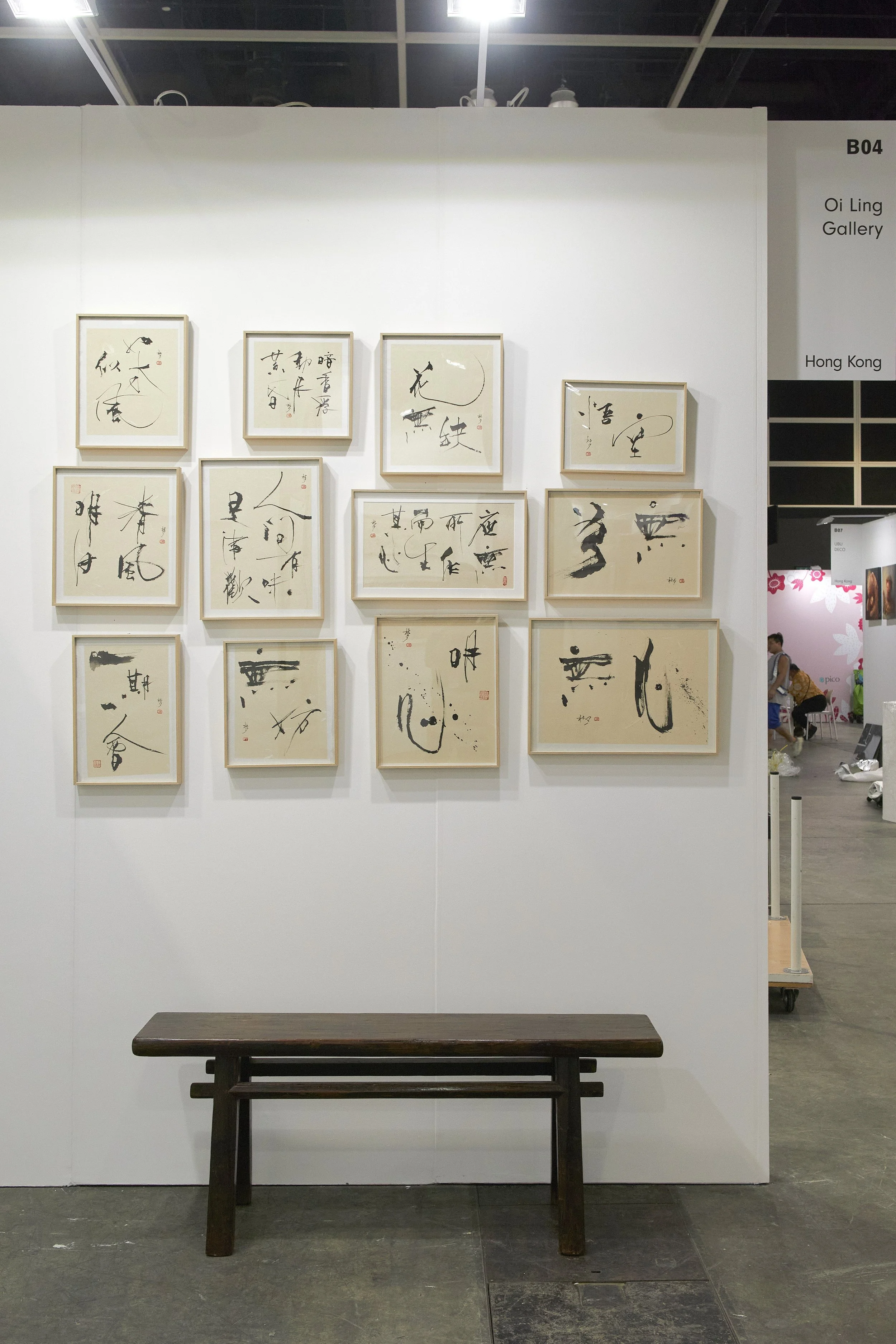 Affordable Art Fair 2025: An art display at an art exhibition featuring framed calligraphy artwork on a white wall with a wooden bench in front, and a sign on the right indicating "Oi Ling Gallery" in Hong Kong.