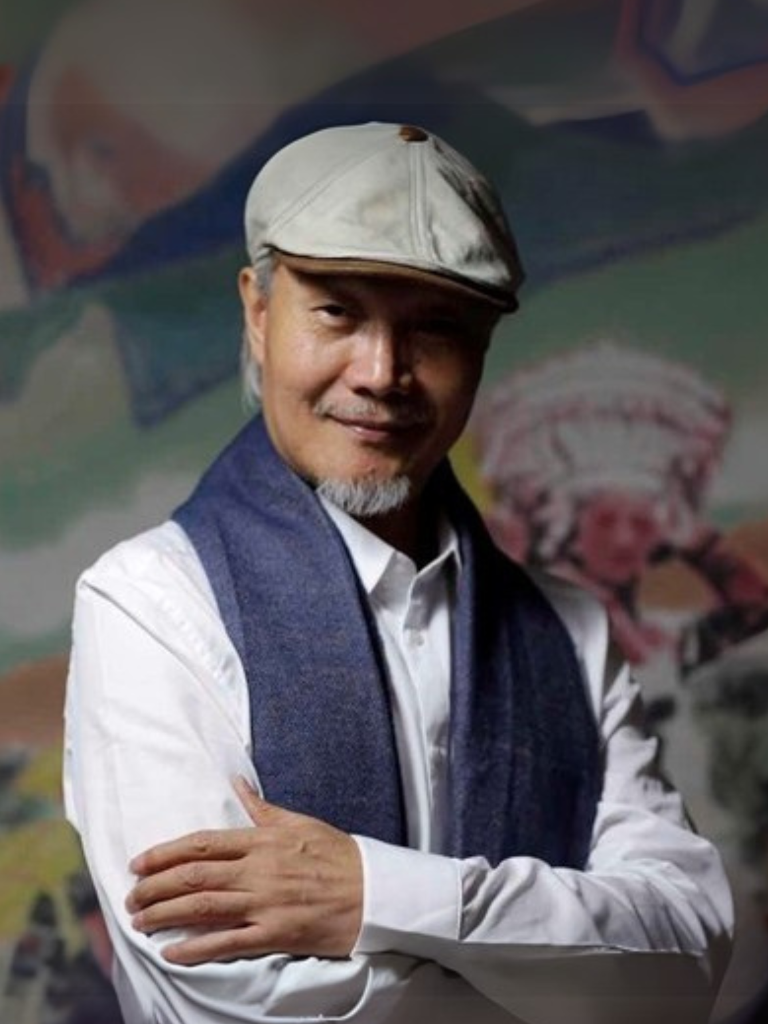 Fan Hongbin 樊鴻賓： A man with gray hair and beard, wearing a white cap, a white shirt, and a blue scarf, smiling with arms crossed in front of a colorful painted mural background.