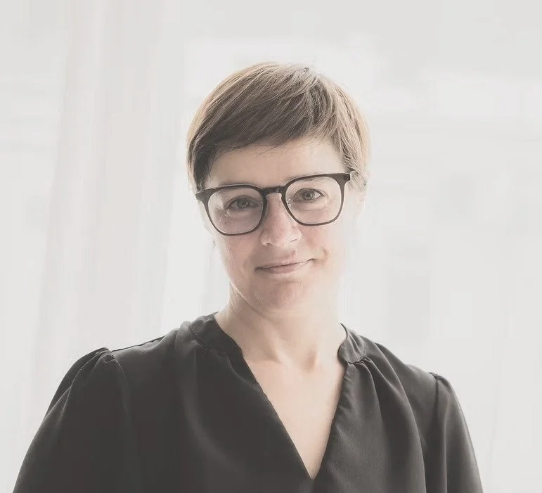 Mia Nel: A woman with short brown hair, glasses, and a black shirt standing indoors with a neutral expression.