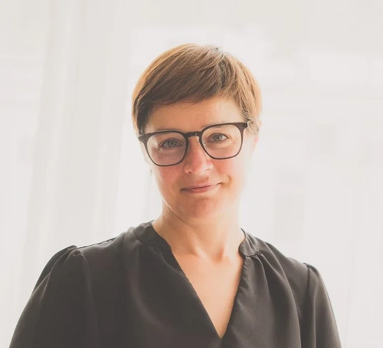 Mia Nel: A woman with short brown hair, wearing glasses and a black top, standing indoors with a bright window in the background.