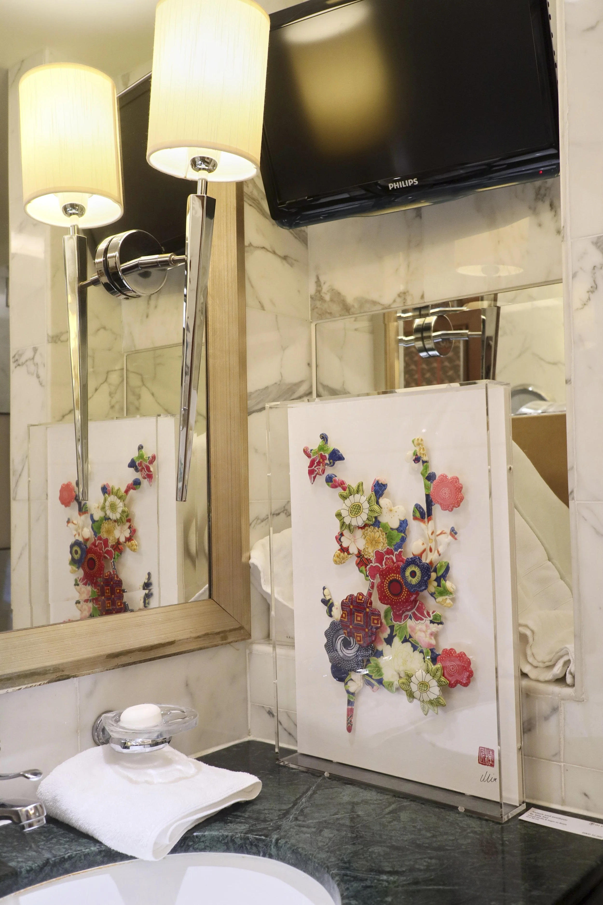 Whatz Art Fair 2025: Decorative artwork featuring colorful floral textures on a white background, placed on a bathroom counter with a mirror and a TV above.